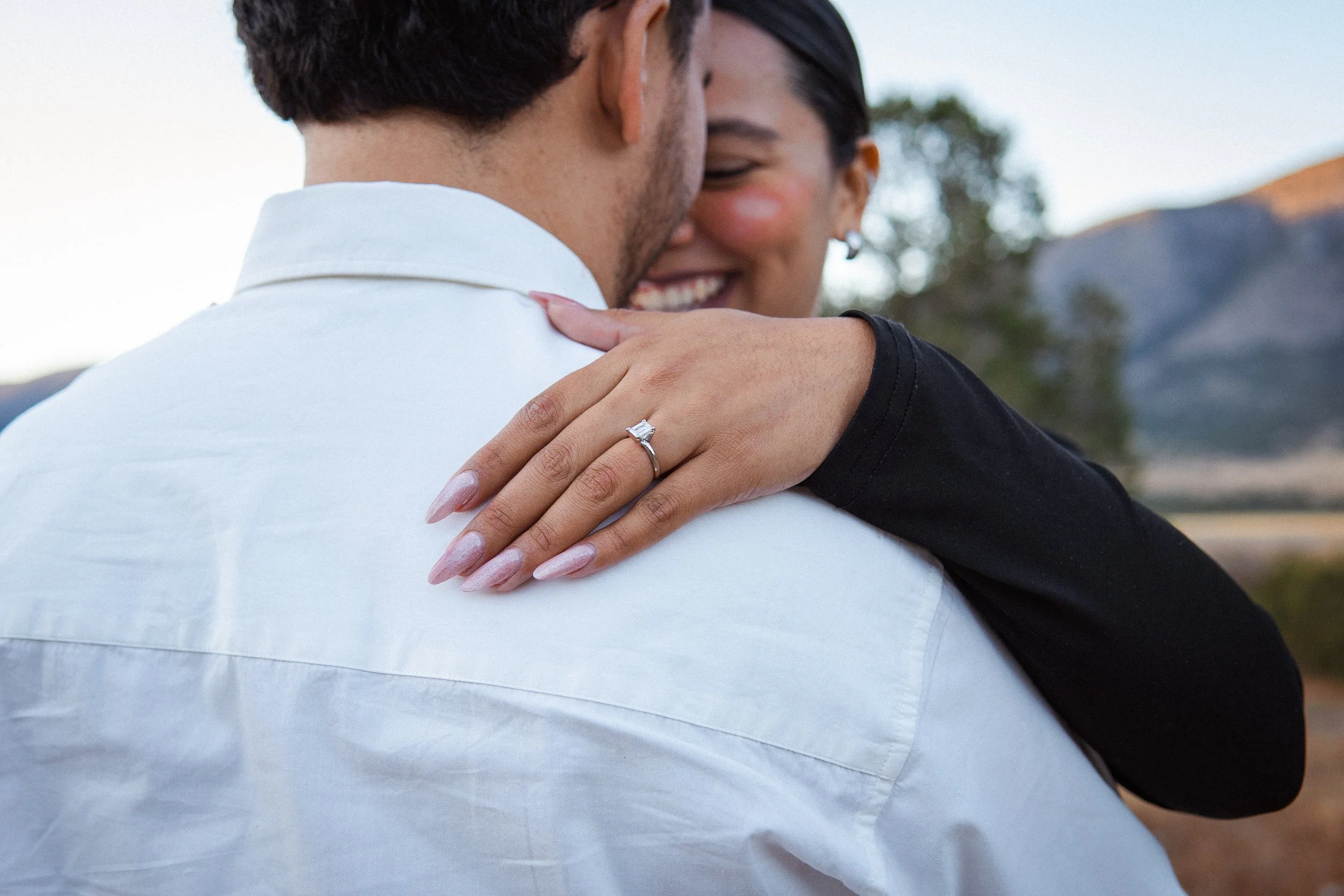 Una pareja abrazándose y sonriendo, con el enfoque en la mano de la mujer que muestra un anillo de compromiso.