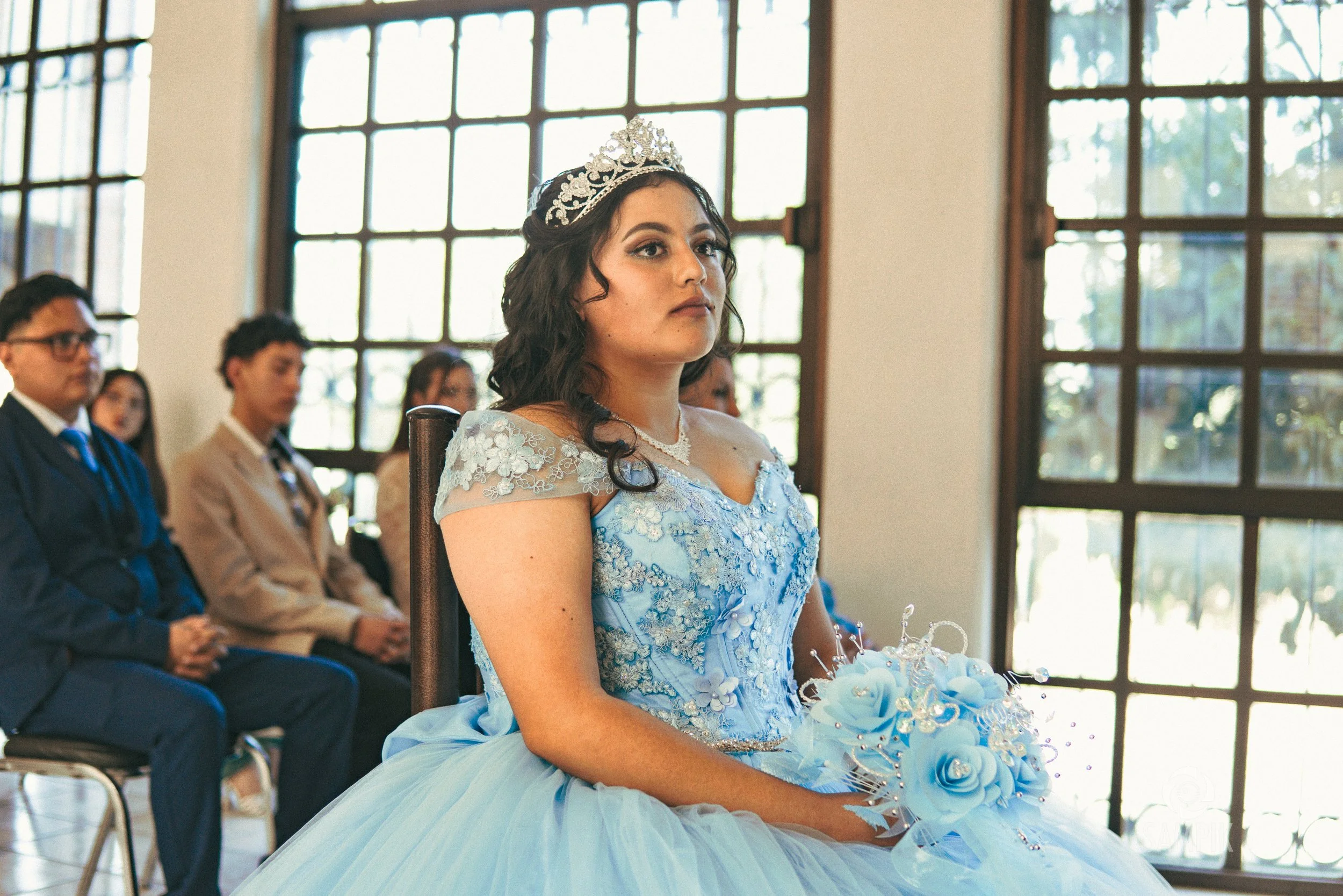 Mujer con vestido de quinceañera azul y corona en una ceremonia, rodeada de personas sentadas y grandes ventanas con luz natural.