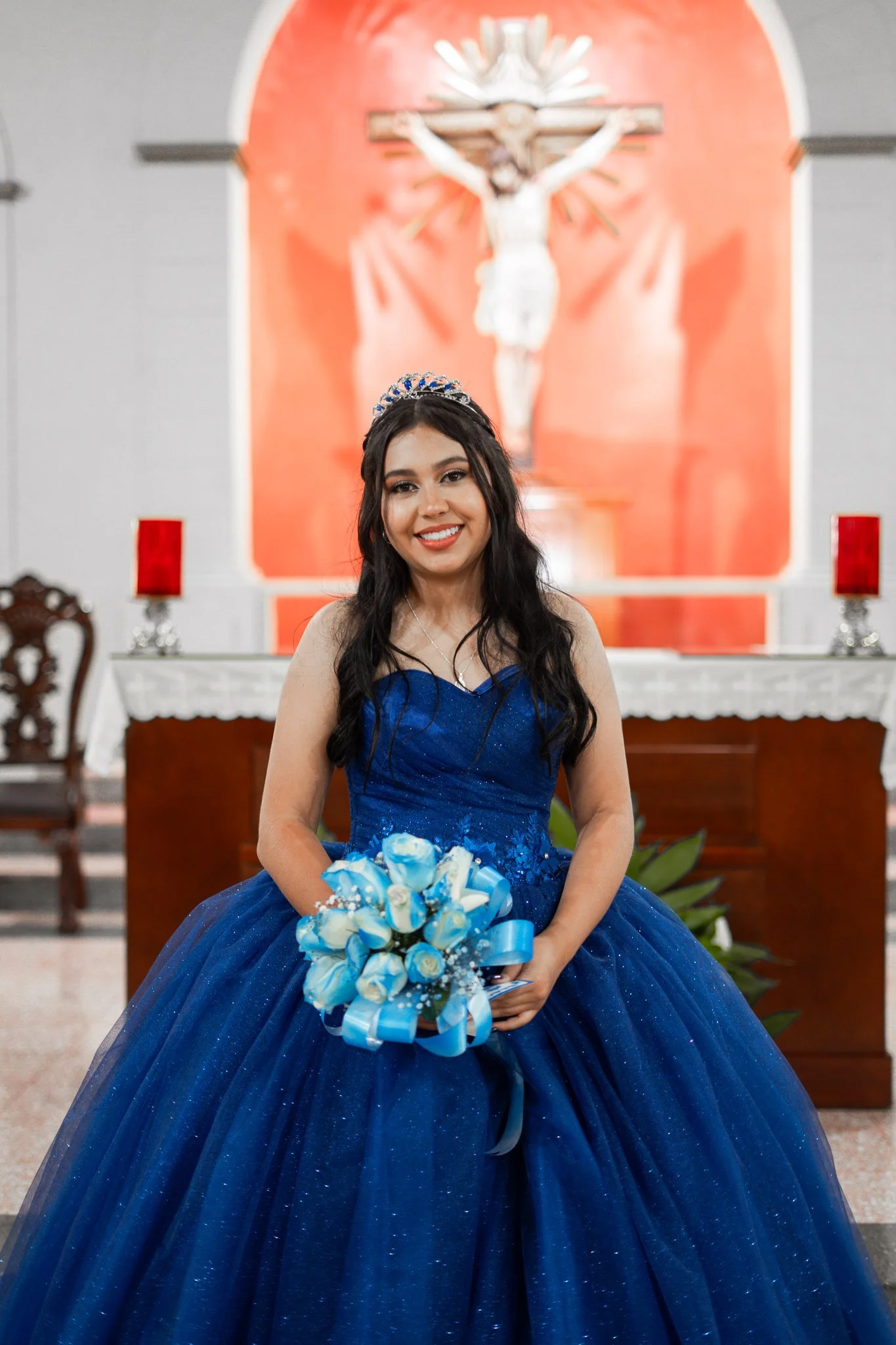 Joven con vestido azul y coronita, sosteniendo un ramo de flores, en una iglesia con altar y crucifijo al fondo.