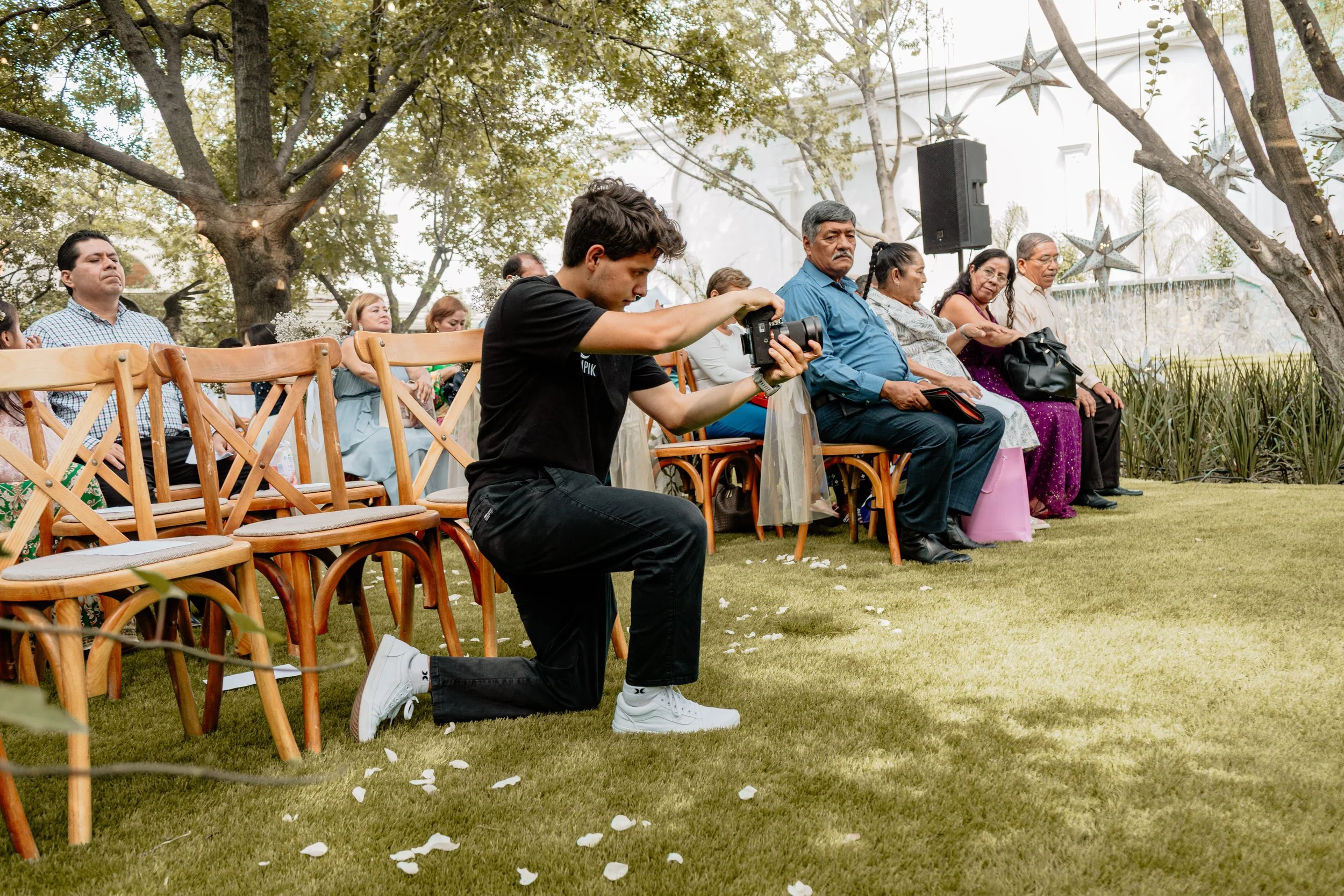 Un grupo de personas sentado en sillas de madera en un evento al aire libre, con un joven tomando fotos con una cámara, en un escenario decorado con estrellas colgantes y árboles.