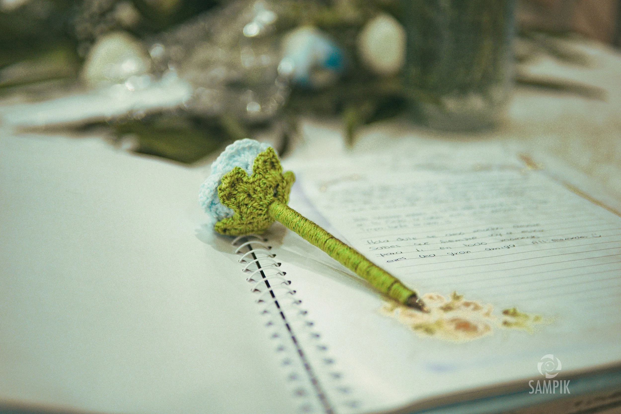 Un cuaderno abierto con una nota escrita a mano, y encima una pluma decorada con hilo verde y una flor de shisa. En el fondo hay objetos borrosos.