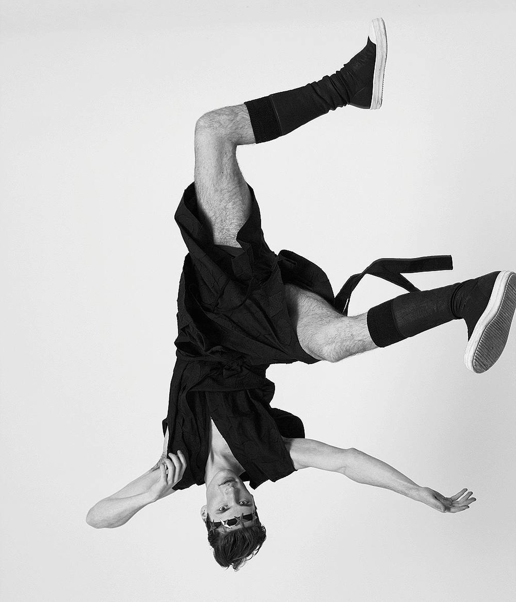 A person is upside down, performing a handstand, with one arm and one leg on the ground and the other arm and leg extended upward. They are wearing a cap, black clothing, and sneakers, against a plain background.