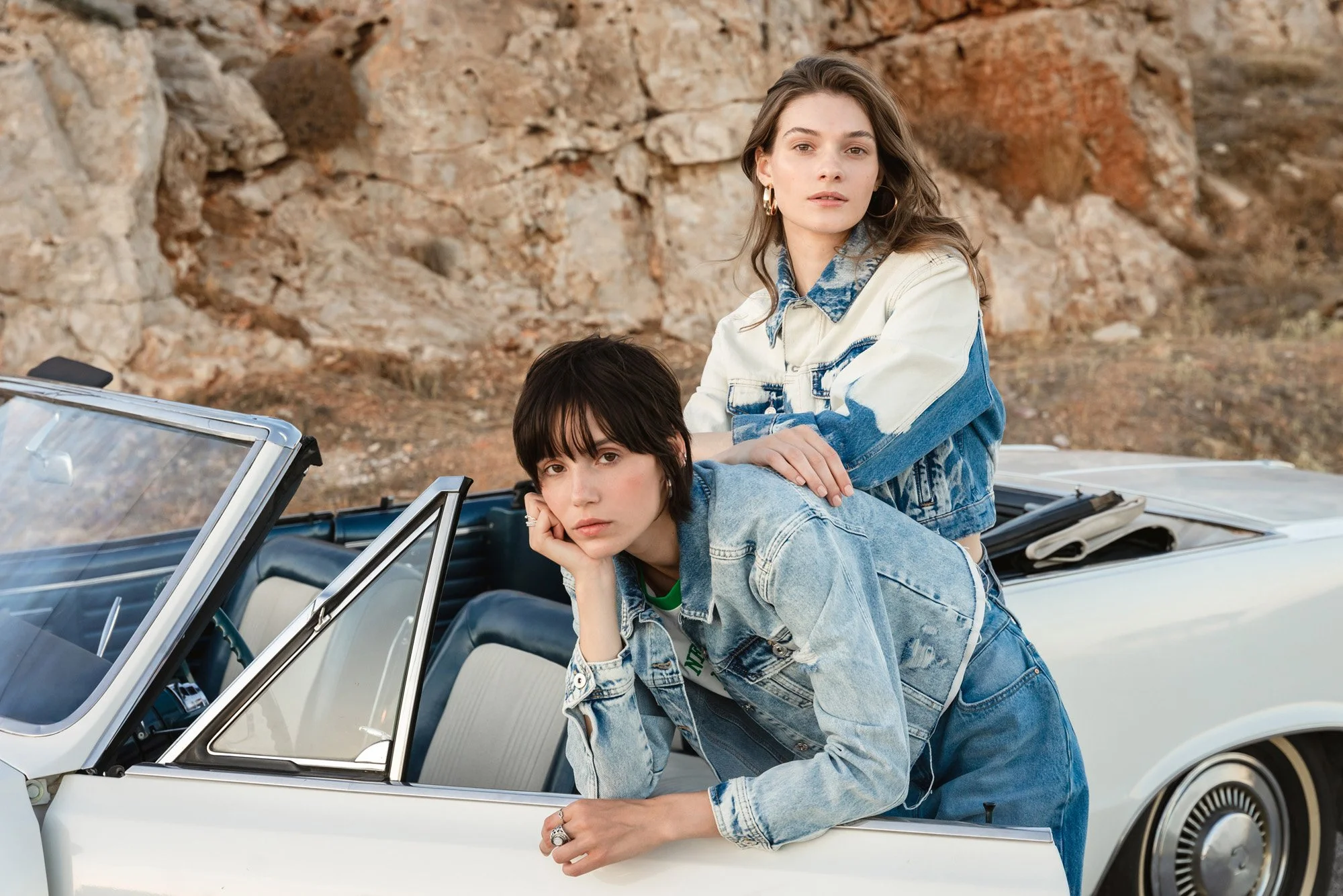 Two young women with casual denim outfits posing on a vintage convertible car, set against rocky desert landscape.