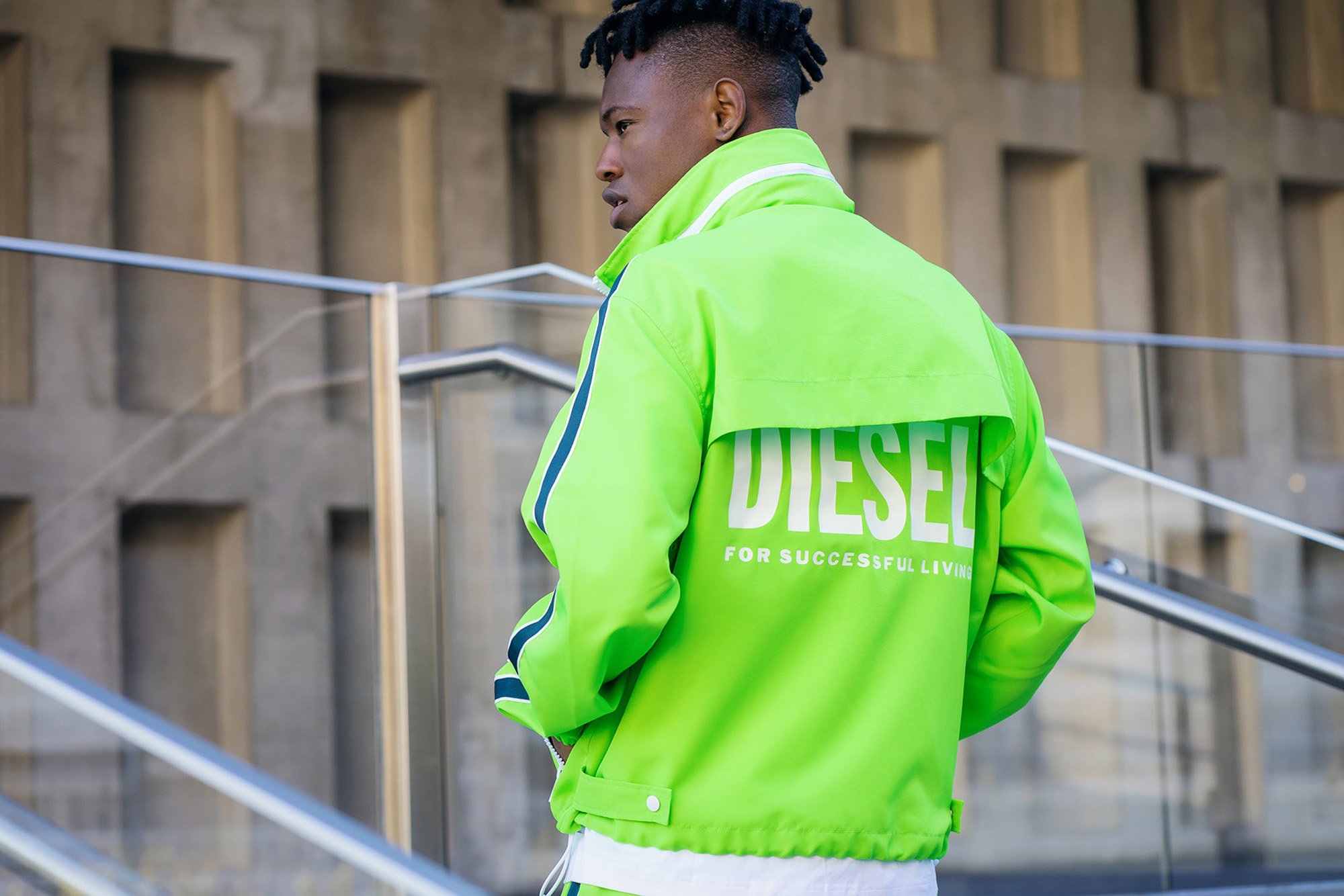 A person with styled dreadlocks wearing a neon green Diesel jacket with the slogan "For Successful Living" on the back, standing outdoors near a modern concrete building with square windows and metal railings.