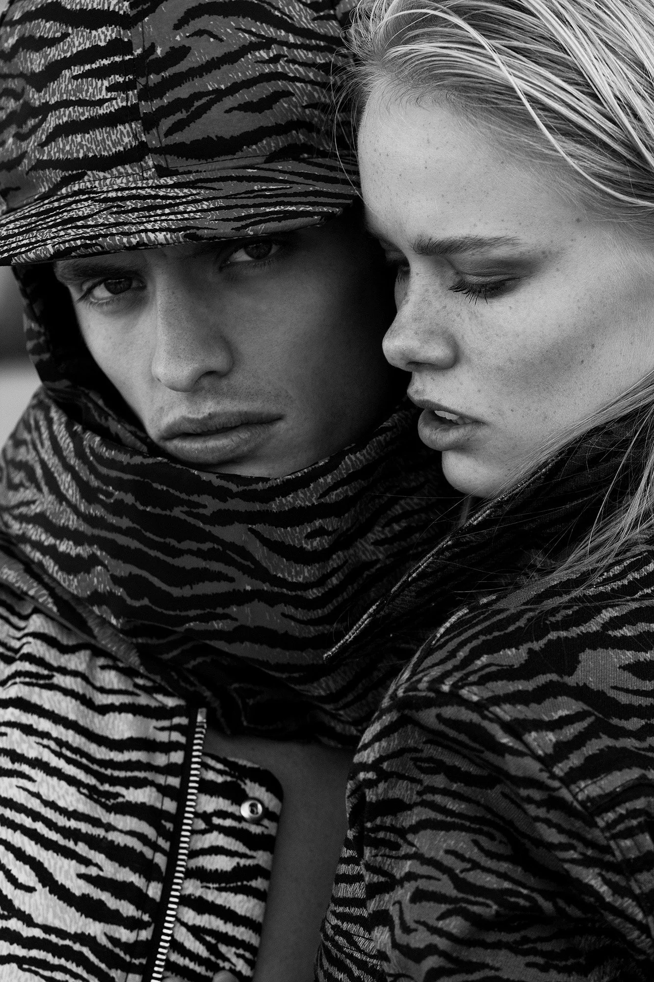 Close-up black and white photo of a young woman and a young man in an intimate pose, with their faces close together. The woman has light hair and freckles, with a serious expression, while the man is wearing a patterned hooded jacket with the hood u