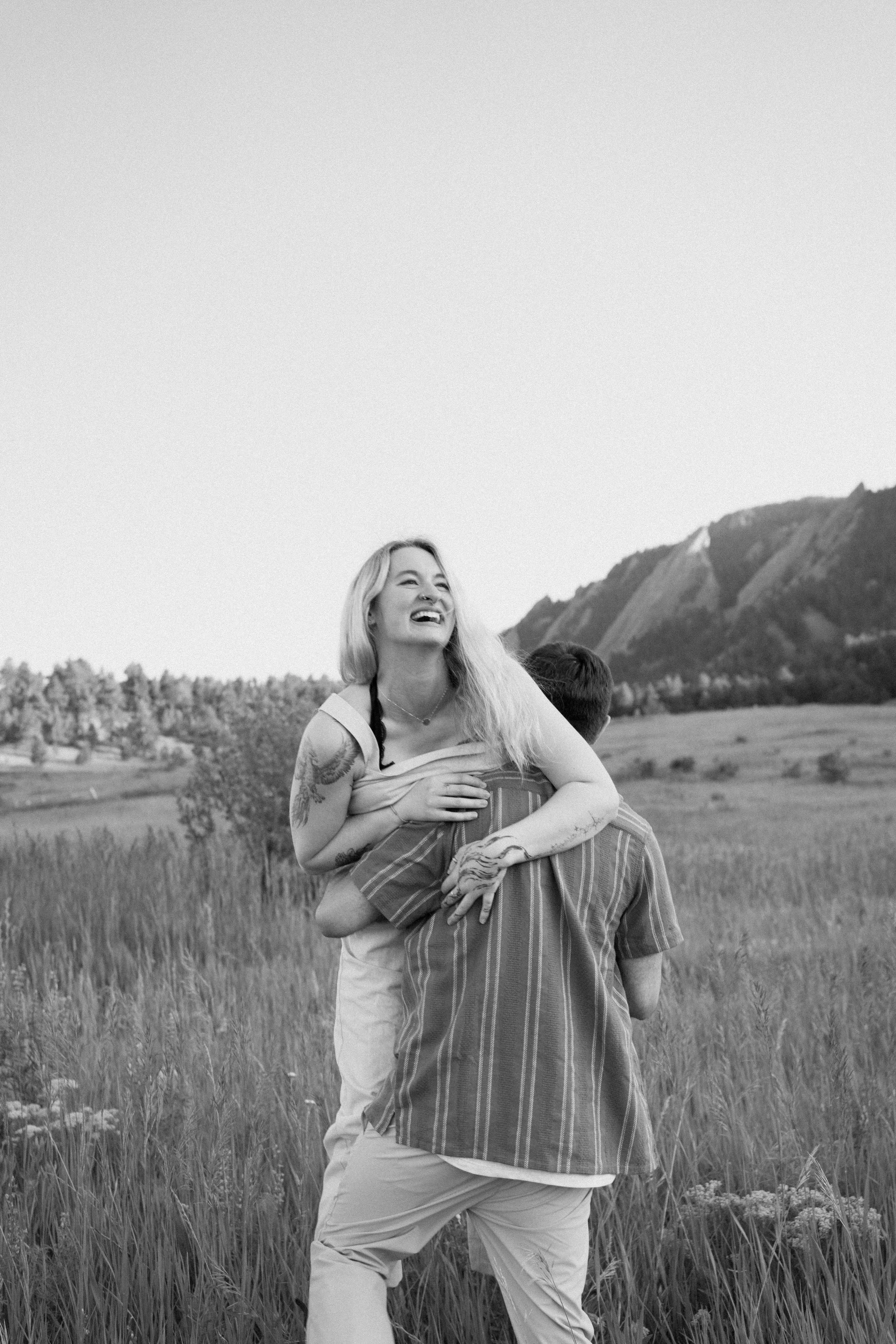 A woman with long blonde hair, smiling and laughing, is being carried on the shoulders of a man in a striped shirt in a grassy field with mountains in the background.