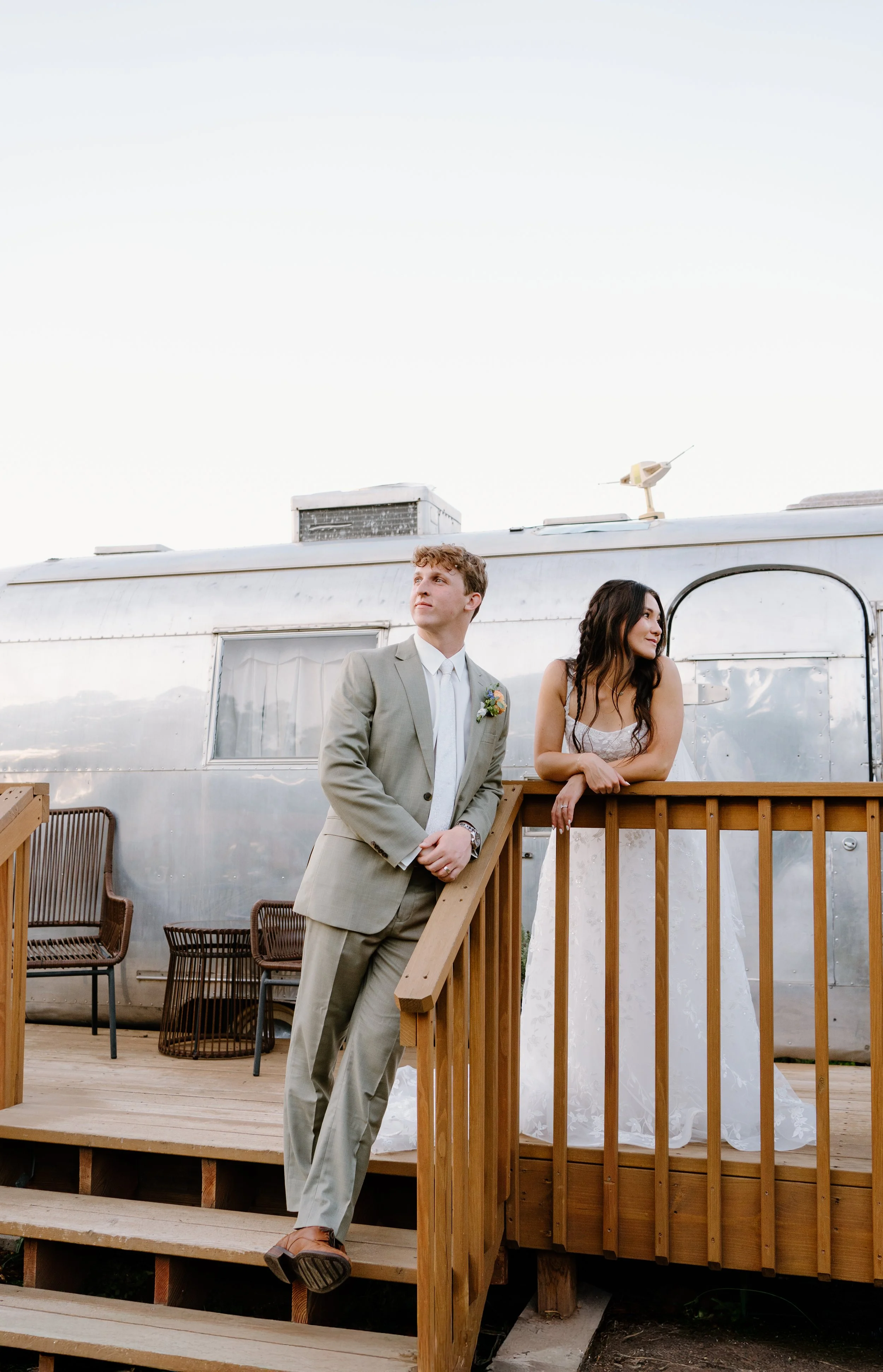 A man in a light gray suit and a woman in a wedding dress stand on a wooden deck outside, leaning on a wooden railing with a metallic trailer or mobile home in the background.