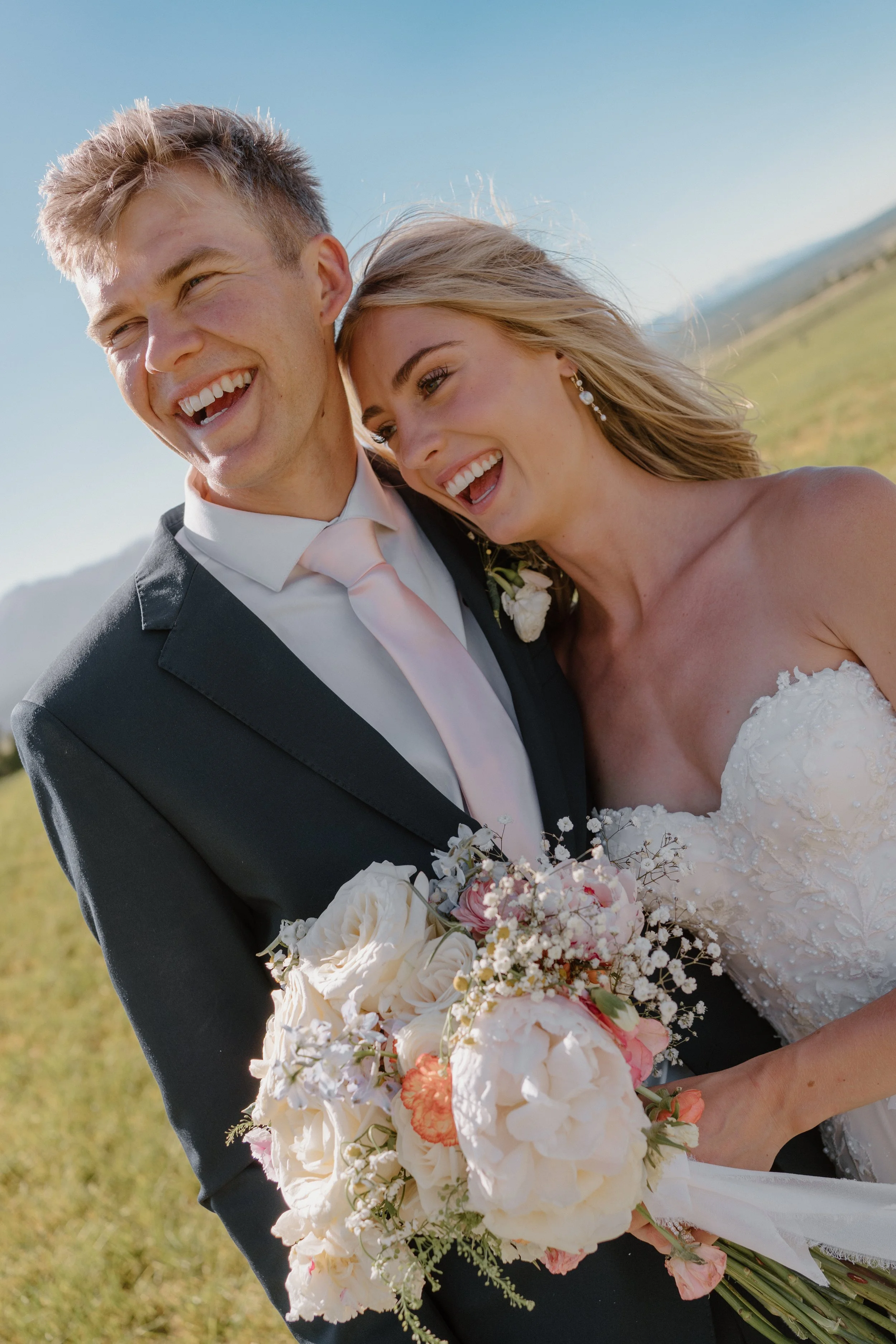 A bride and groom smiling and embracing outdoors, holding a bouquet of white and pink flowers, with a scenic field and blue sky in the background.