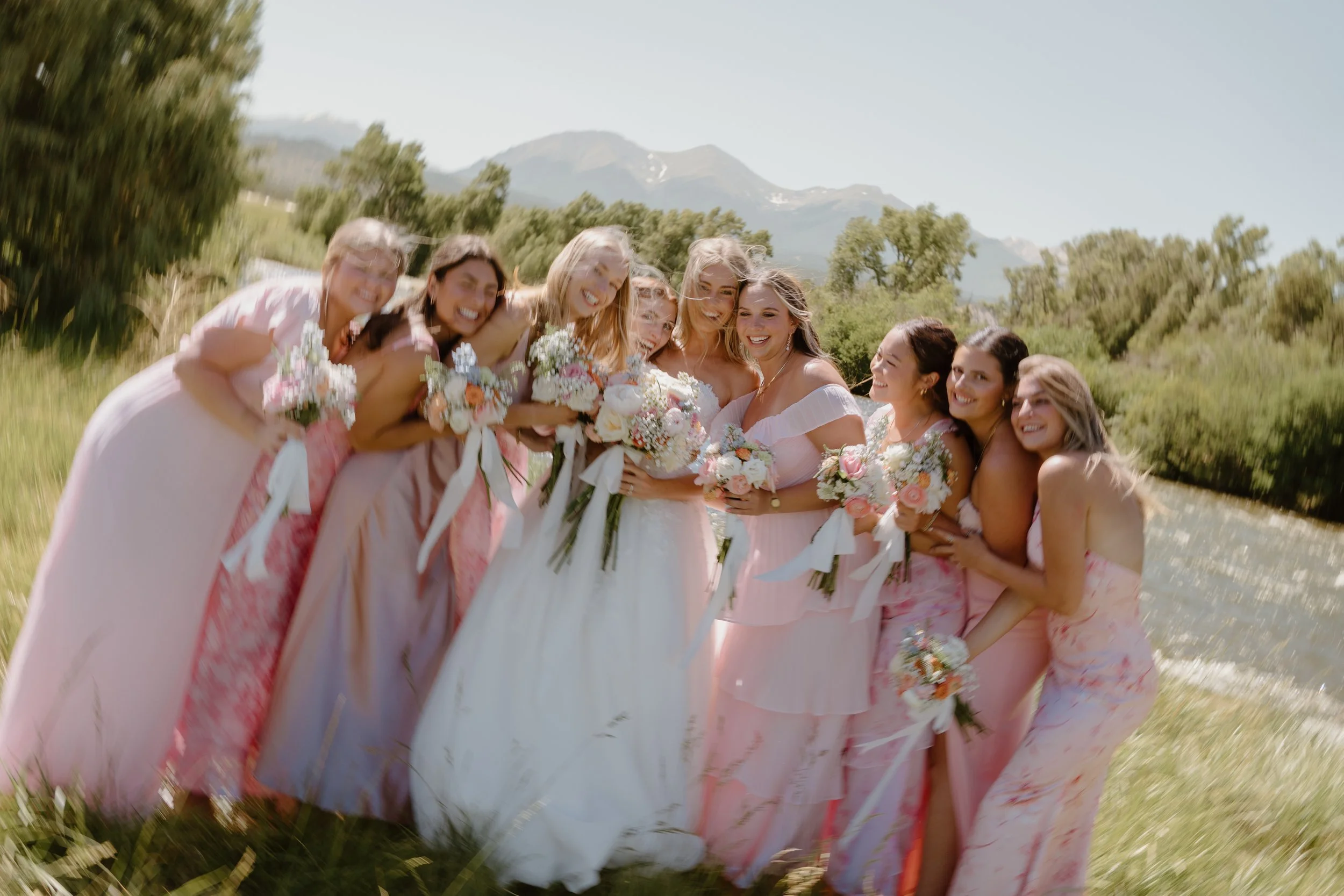 A bride and her bridesmaids standing outdoors in a field with mountains in the background, smiling and holding bouquets of flowers.