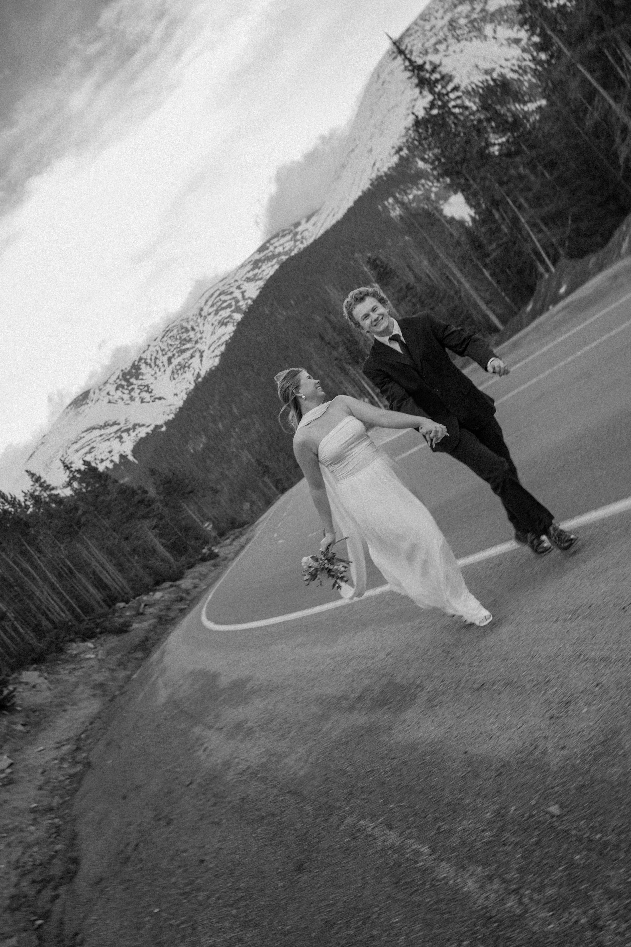A black and white photo of a newlywed couple holding hands and walking across the road in a mountainous landscape with snow-capped peaks and tall pine trees.