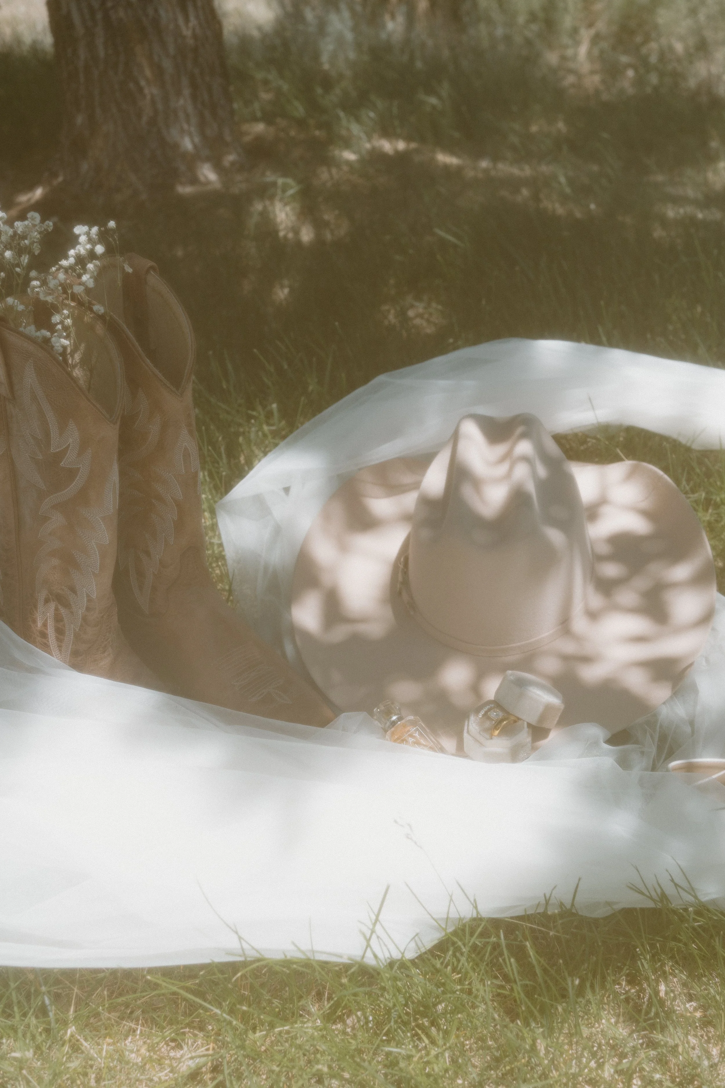 A woman lying on the grass in a sunny outdoor setting, wearing a large sunhat, with a white veil or tulle misted over her. She is surrounded by shadows of tree leaves and is wearing cowboy boots.