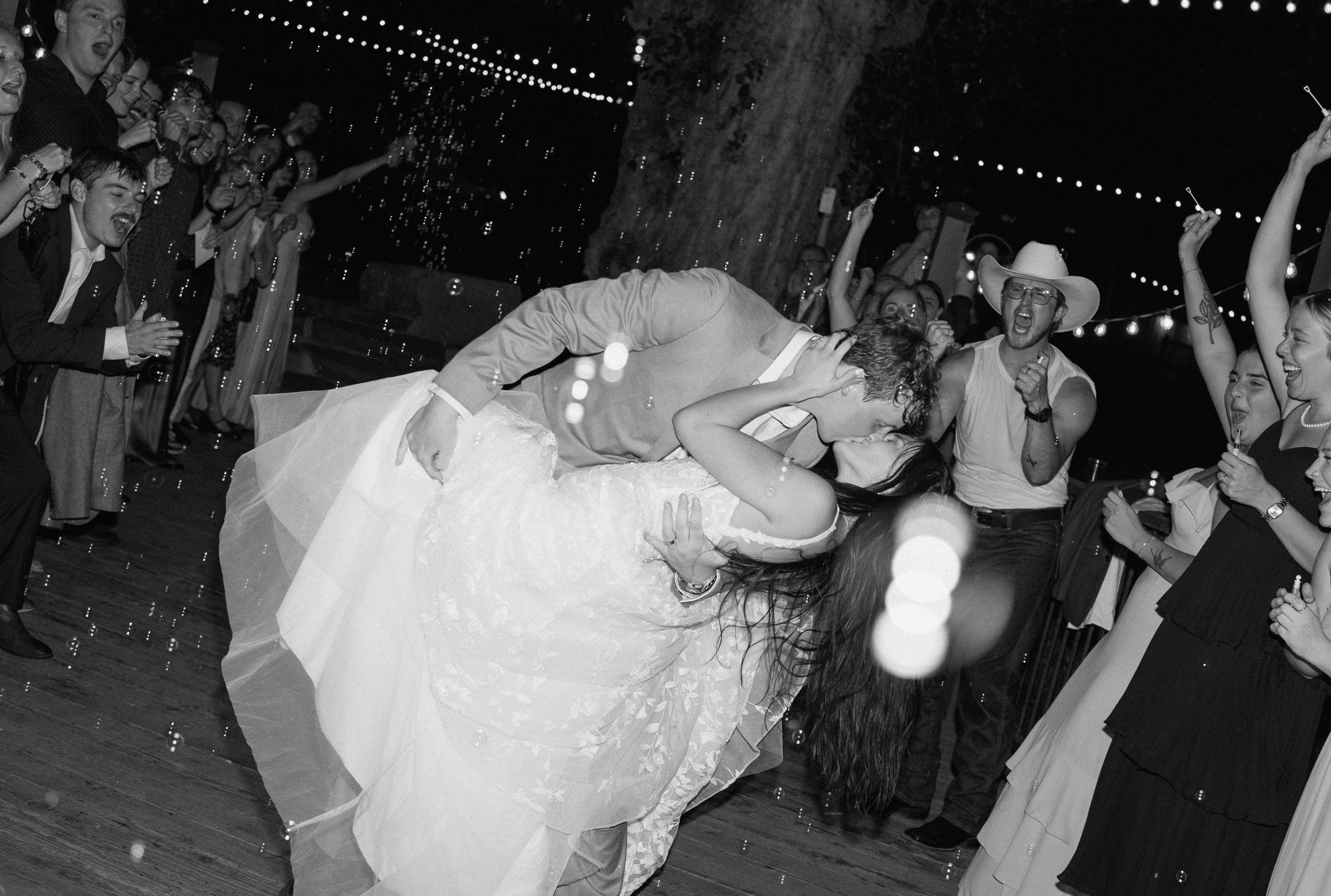 A bride and groom kissing during their wedding exit with guests surrounding them, some cheering and clapping.