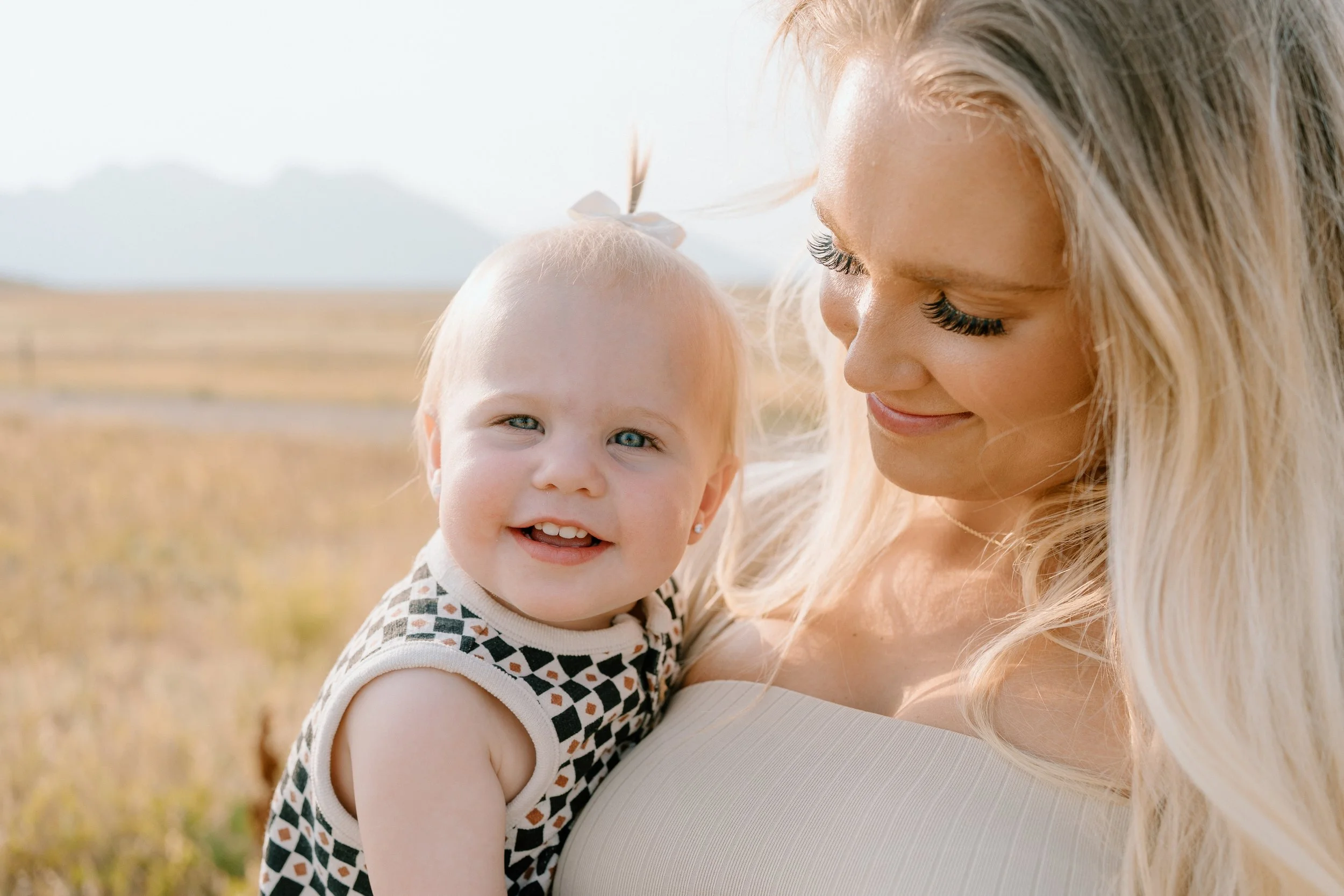 A smiling woman holding a happy toddler girl outdoors in a field with mountains in the background.