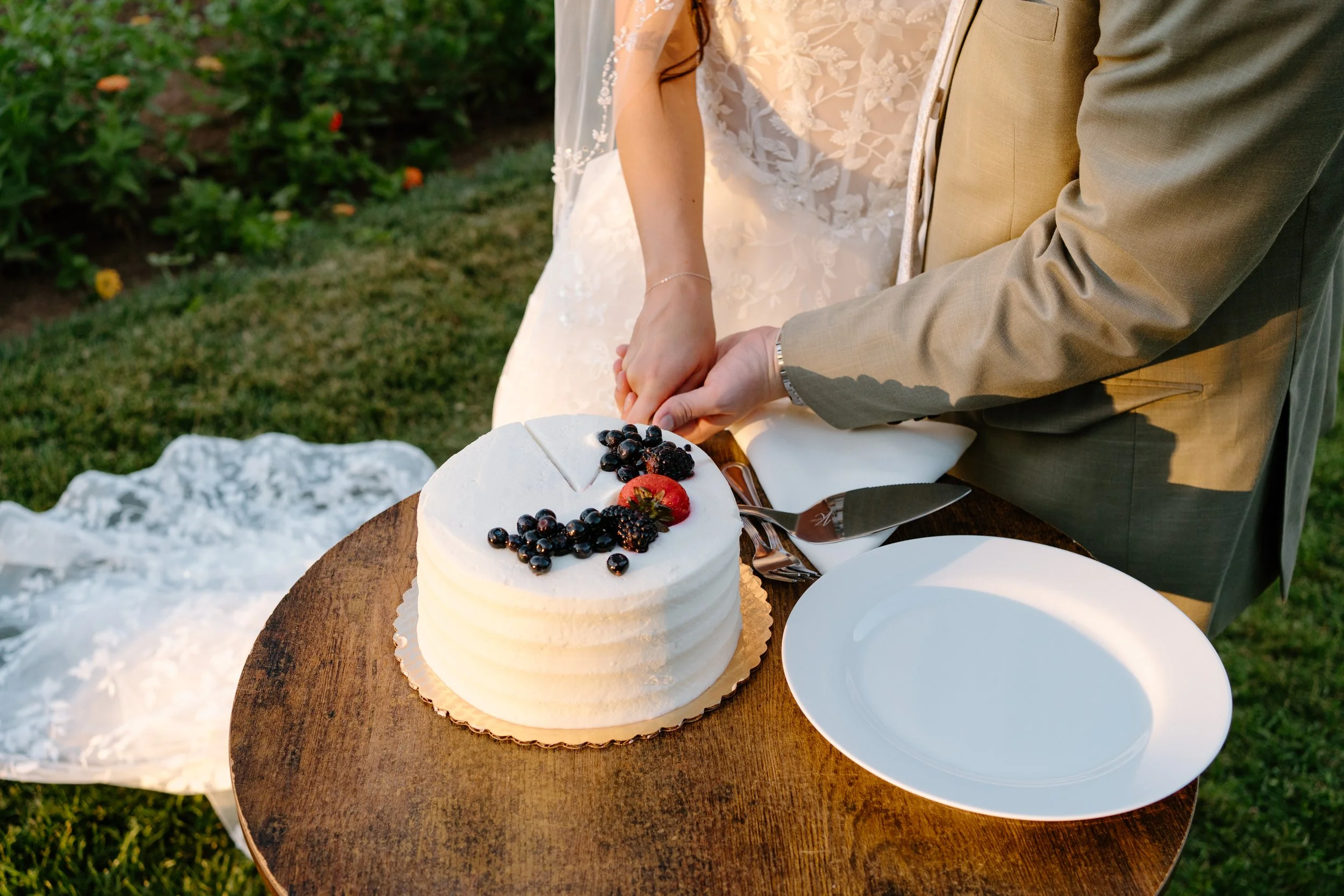 A newlywed couple cutting a white wedding cake topped with blackberries, blueberries, and a strawberry, with a garden in the background.