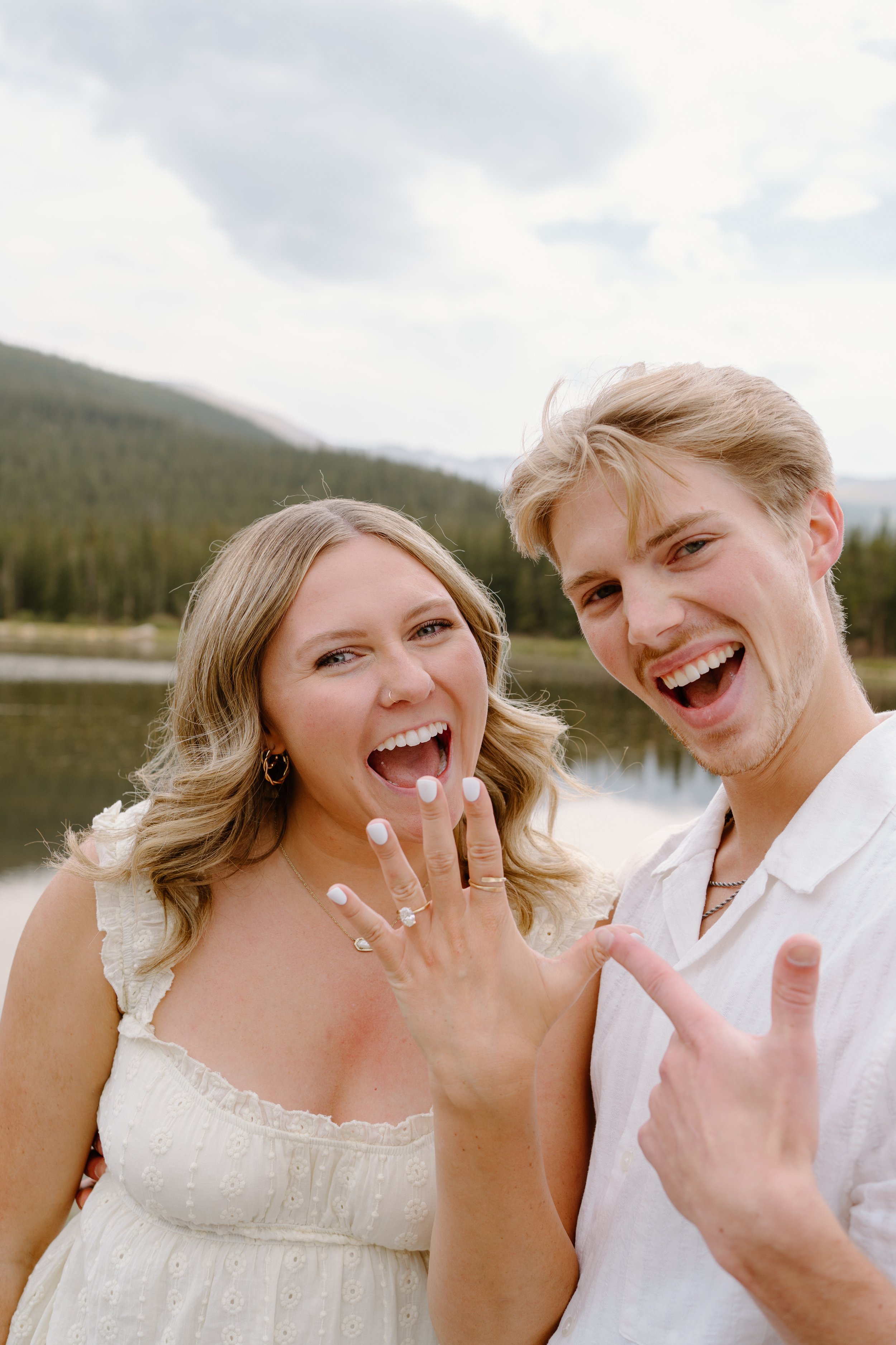 Happy young couple outdoors by a lake with mountains in the background, celebrating with the woman showing her engagement ring.