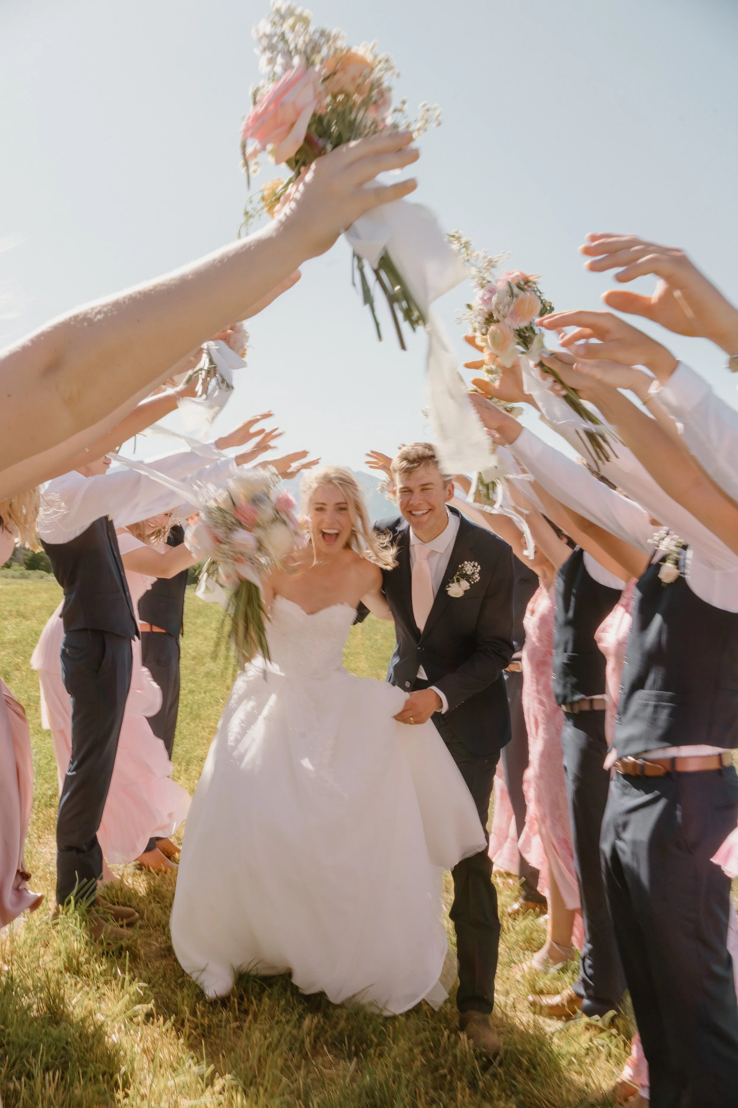 A wedding celebration outdoors with a bride in a white wedding dress and a groom in a black suit walking through a tunnel of guests holding bouquets, with sunny weather and green grass.