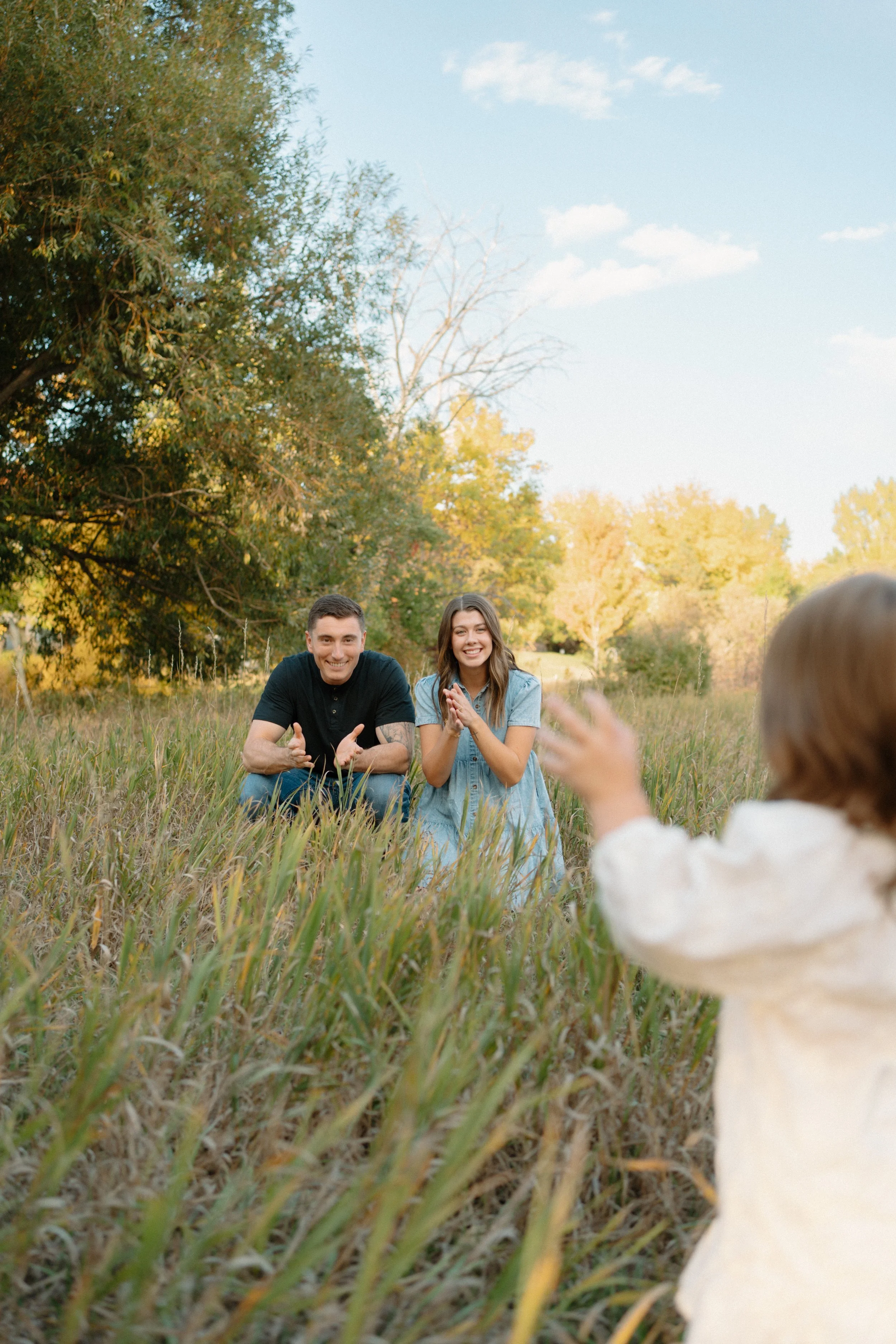A young daughter reaching out her parents who are kneeling in a grassy field outdoors during late afternoon in a mini photography session.