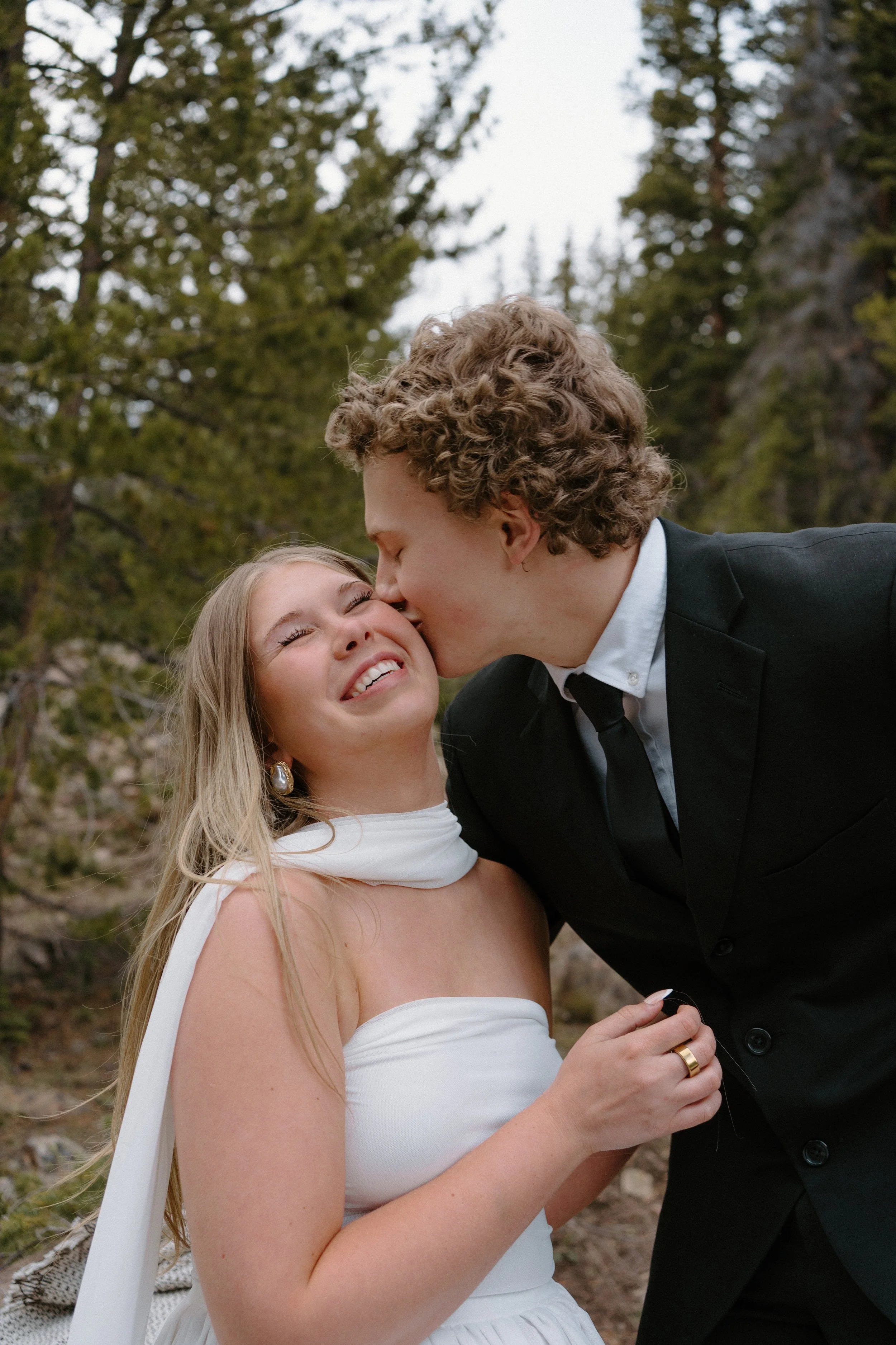 A couple dressed in wedding attire sharing an outdoor kiss, the man wearing a black suit and the woman in a white dress with pearl earrings, in a forested area.