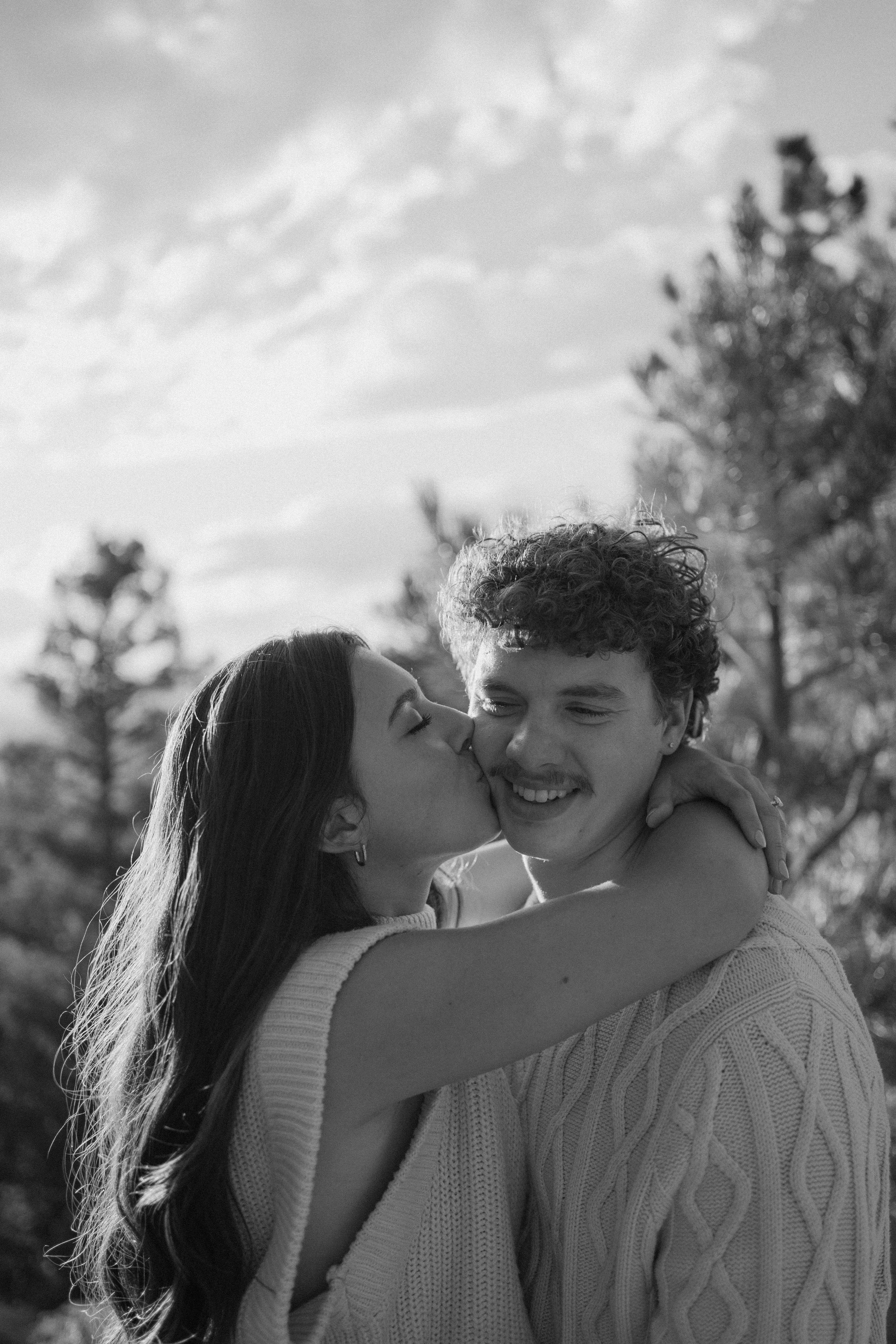 A black and white photo of a woman kissing a man on the cheek while embracing him outdoors with trees and clouds in the background.