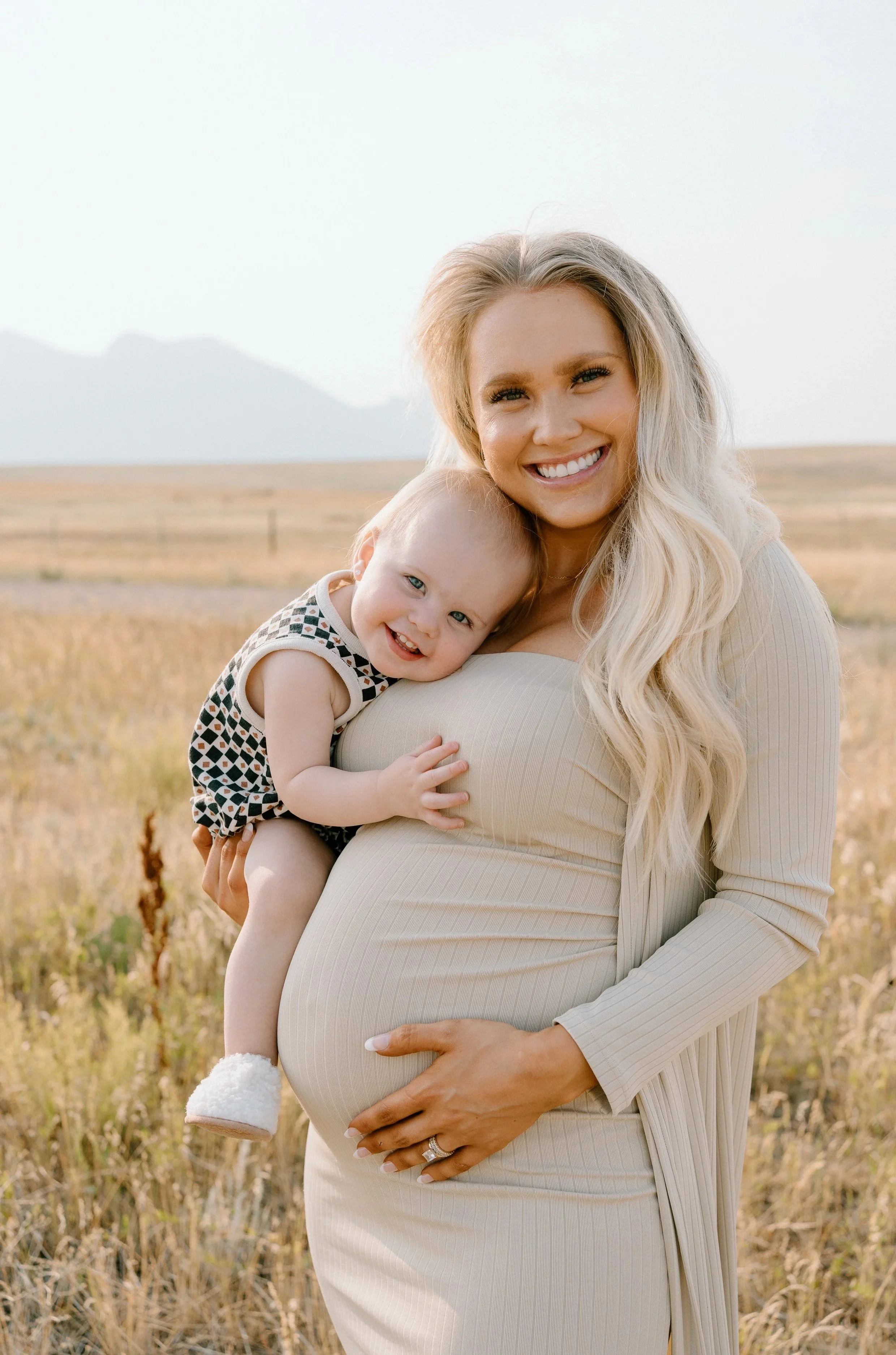 Smiling pregnant woman in beige dress holding young child in a field with mountains in the background in a mini photography session.