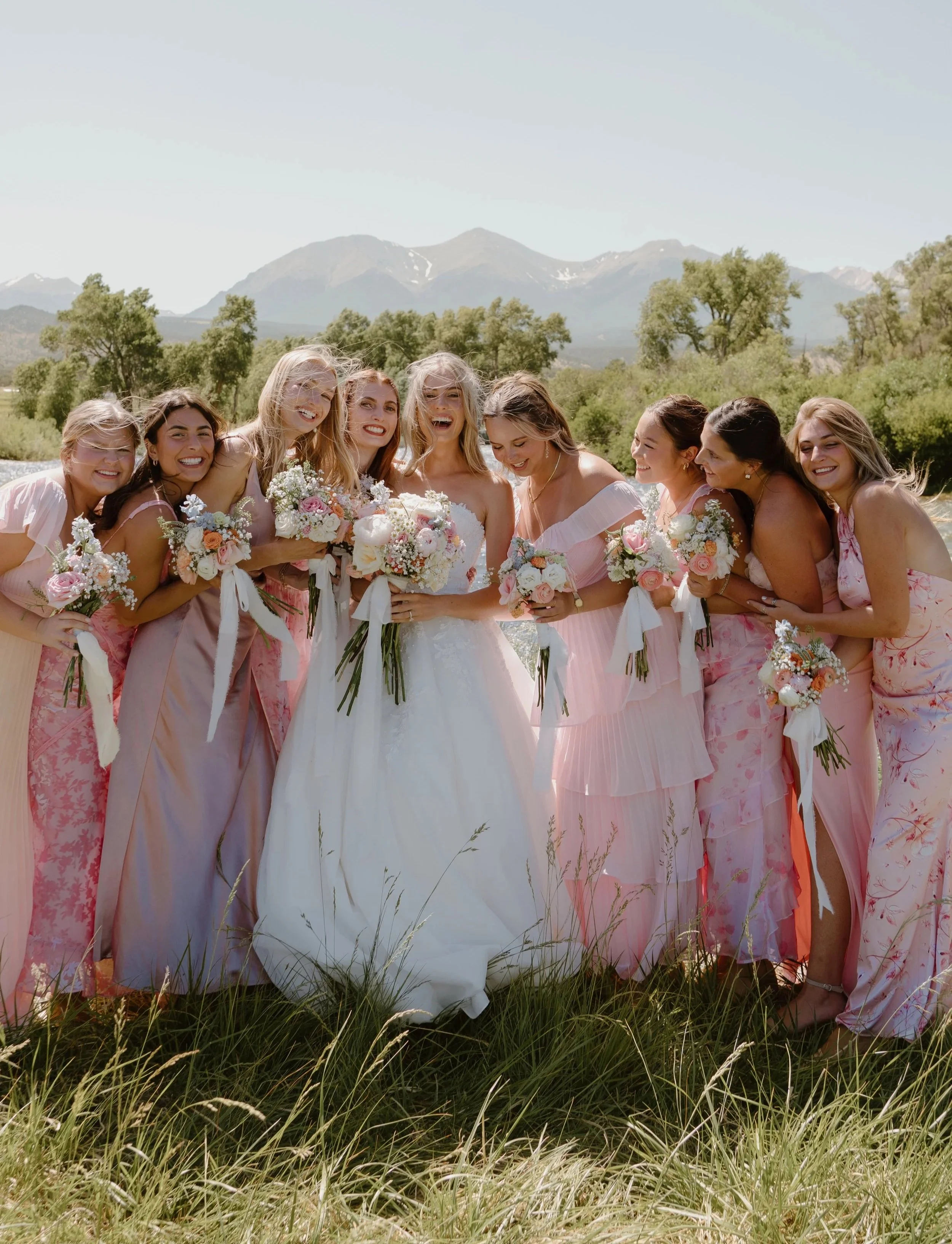 A bride in a white wedding dress surrounded by her bridesmaids in pink dresses, holding bouquets of flowers, outdoors with mountains and greenery in the background.