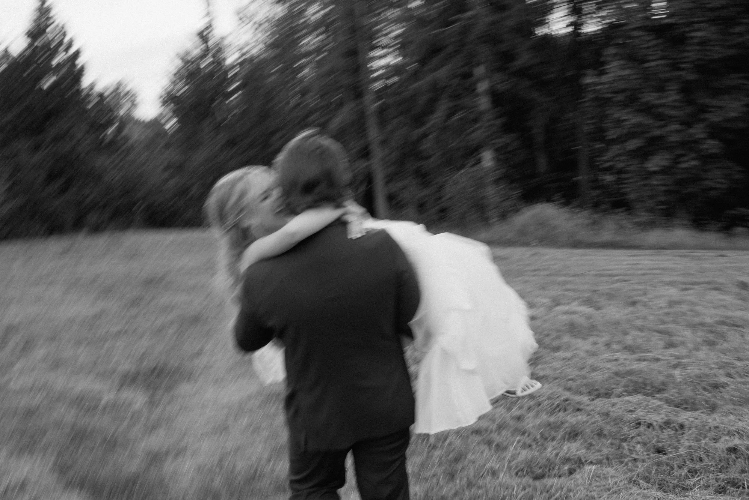 A groom in a suit is carrying his bride in a wedding dress outdoors on a grassy field, with trees in the background, in black and white.