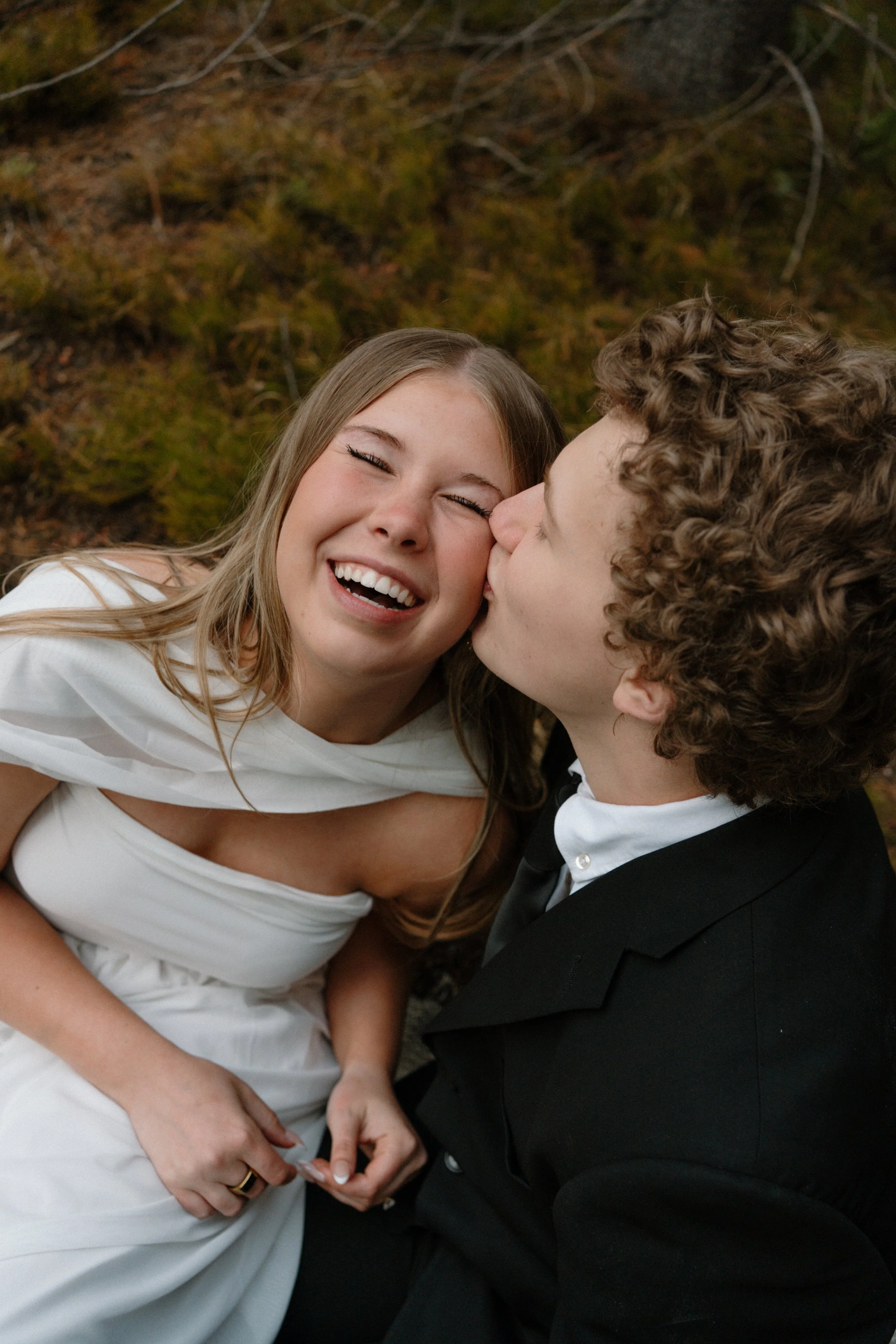 A young couple sharing a kiss outdoors with a mossy ground background, she is smiling and dressed in a white dress, he is in a black suit.