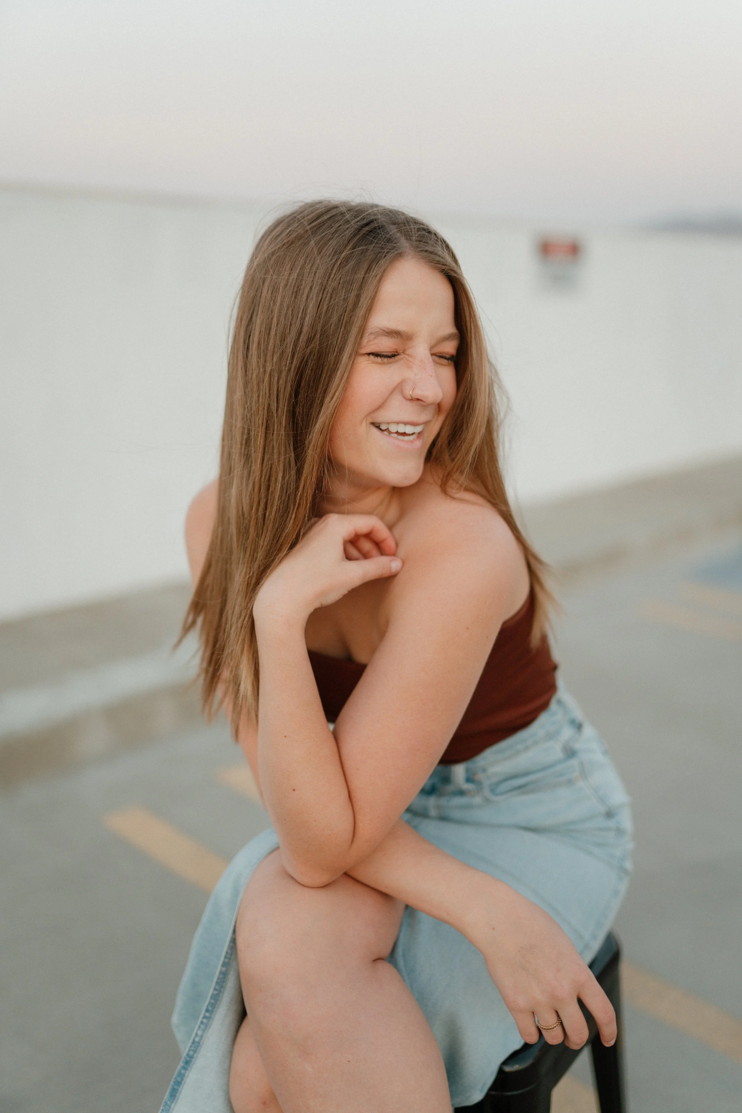 A young woman with long brown hair laughing, sitting on a black chair in a parking lot with a white wall behind her.