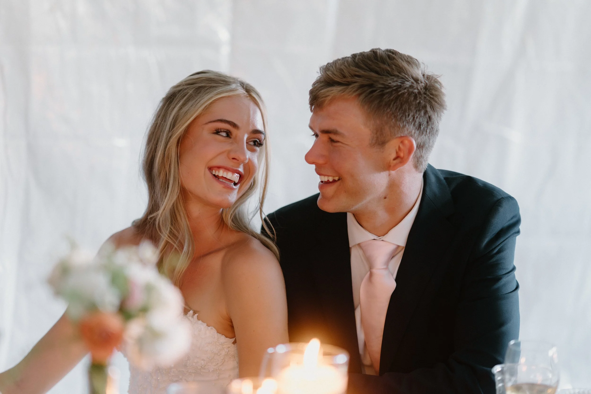 A newlywed couple sitting close together at their wedding reception, smiling and looking at each other affectionately, with a blurred bouquet and a candle in the foreground.