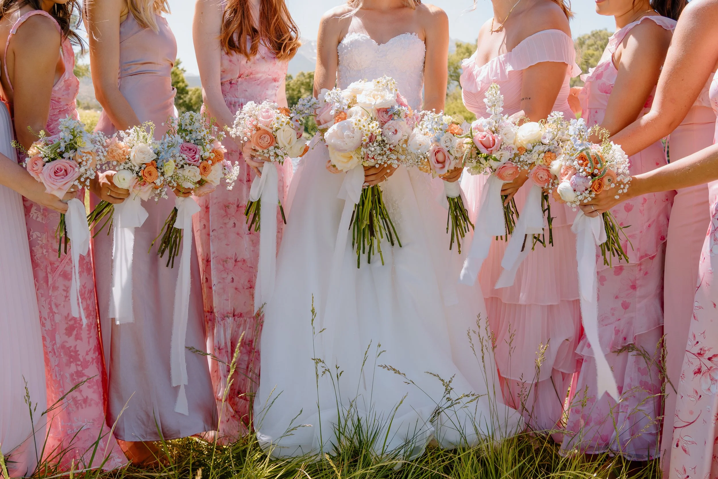 Bride in a white wedding dress holding a bouquet, standing among bridesmaids dressed in pink, holding bouquets, in an outdoor setting.