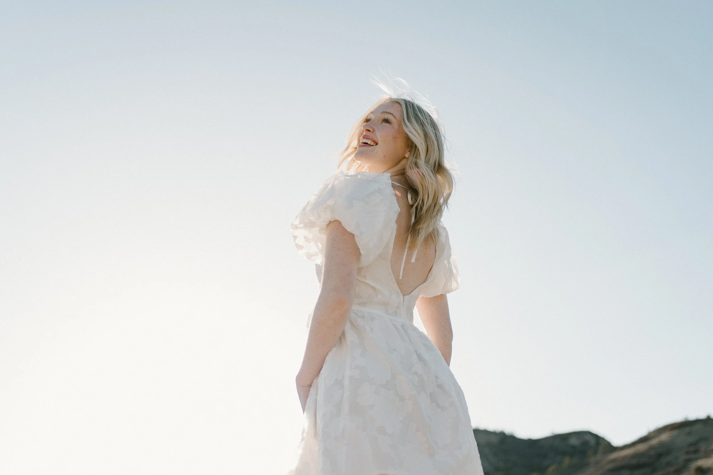 A young woman in a white dress, smiling and looking up, standing outdoors with hills in the background on a clear day.