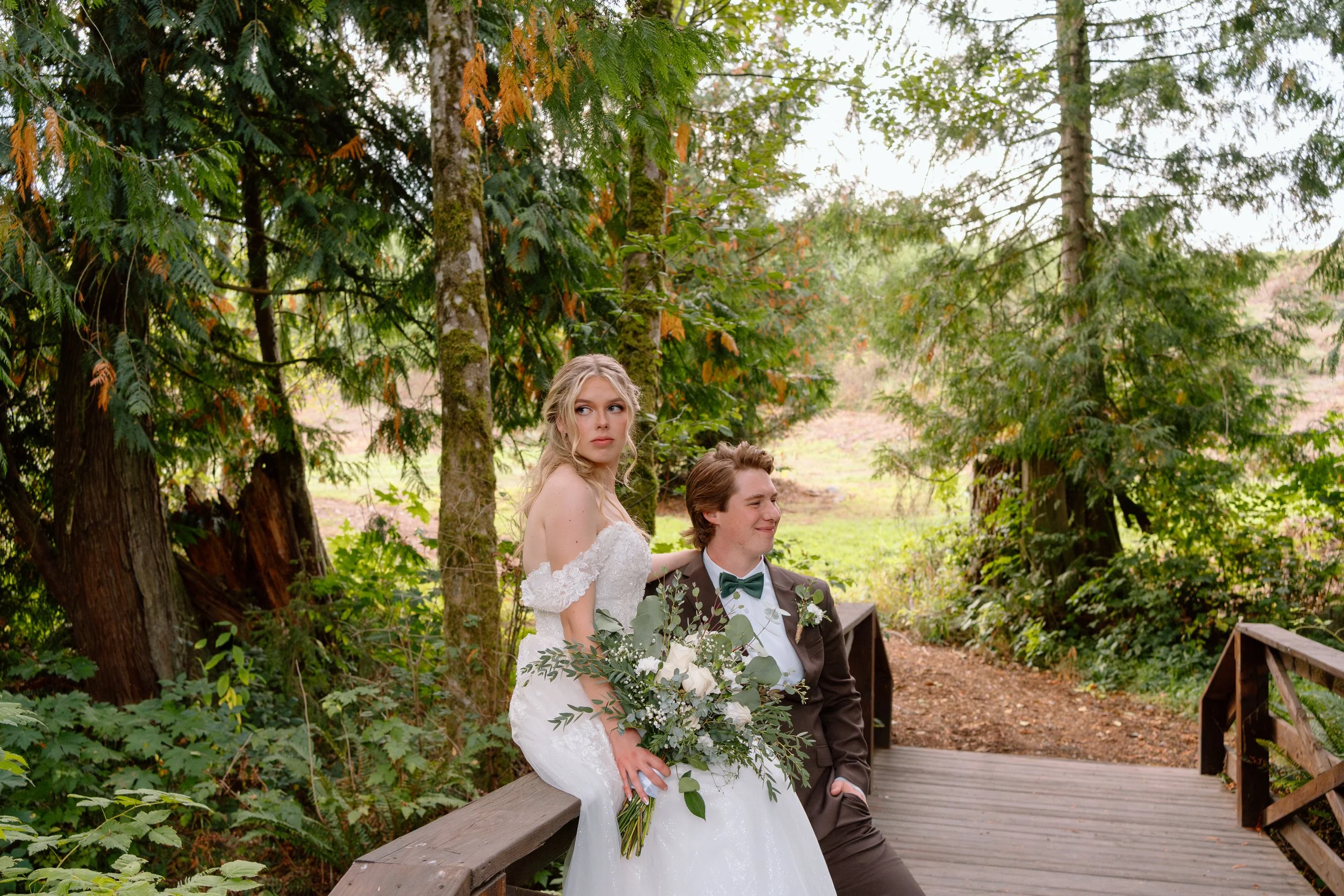 A bride and groom sitting on a wooden bridge in a forested outdoor setting during their wedding in Washington state.
