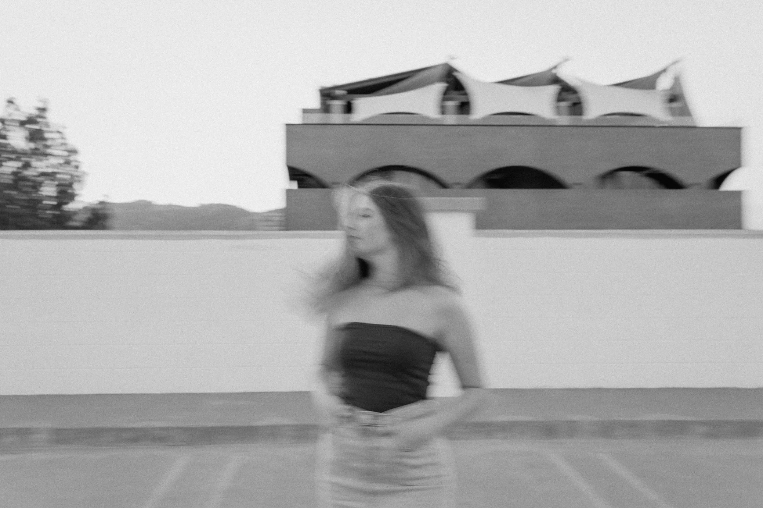 Black and white photo of a woman with long hair in a strapless top and skirt standing outdoors on a rooftop or terrace, with a modern building with arches and a tented roof behind her.