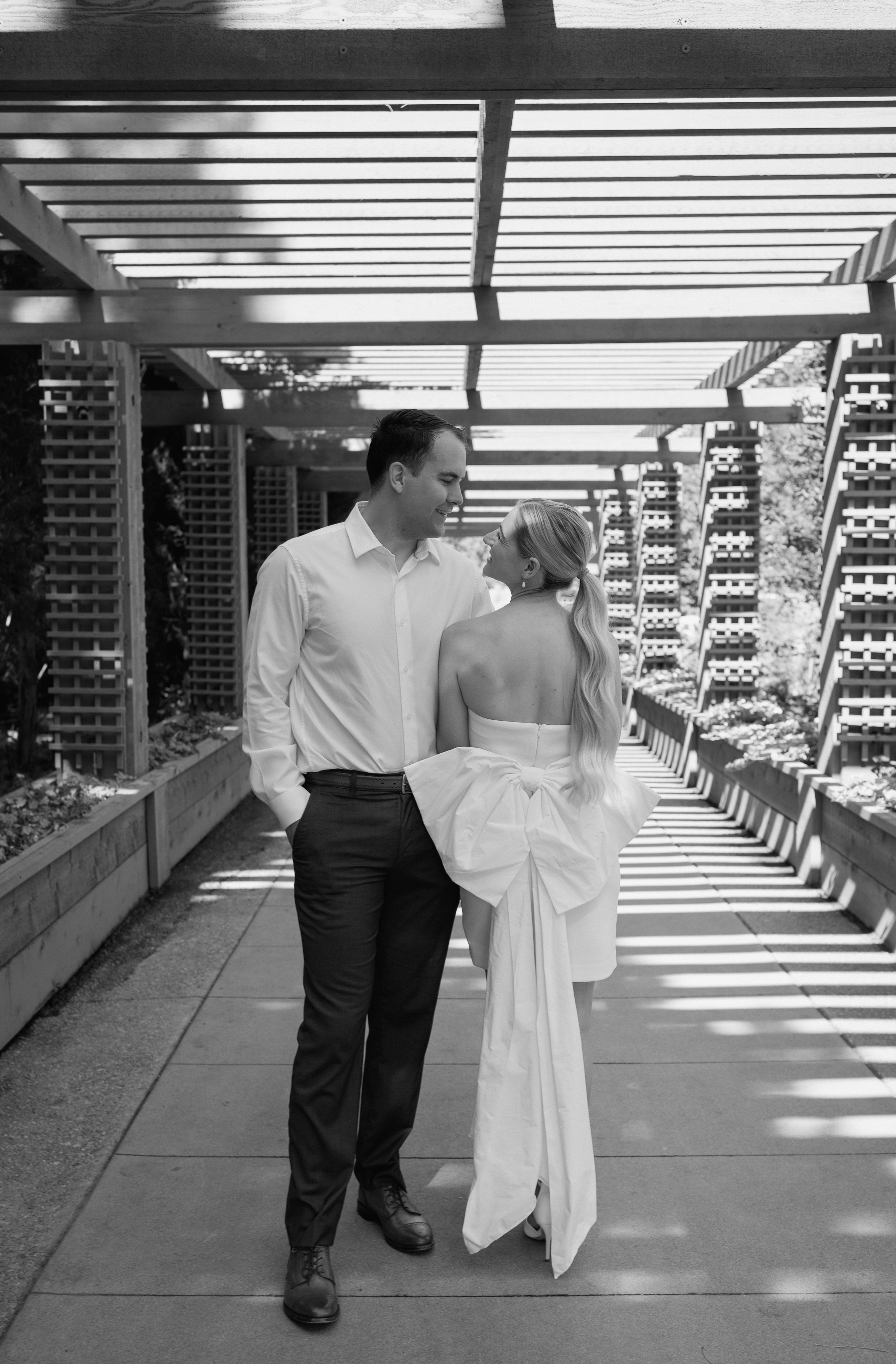 Black and white photo of a couple in formal attire sharing an intimate moment on a walkway with modern architectural structures overhead.