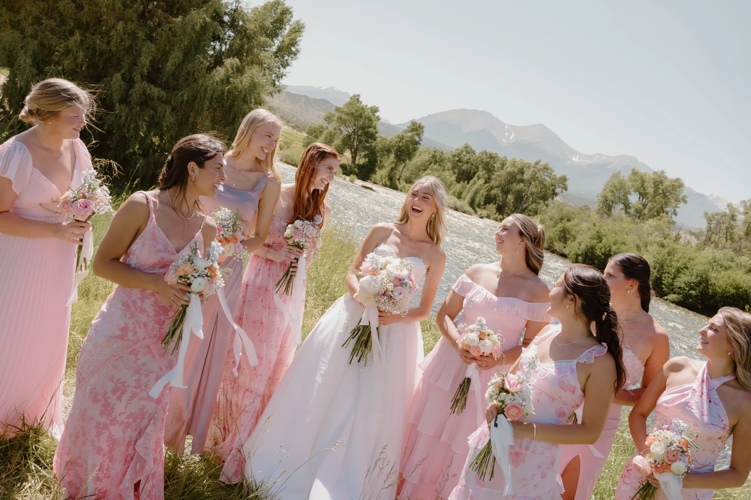Bride laughing with her bridesmaids in soft pink dresses during an outdoor wedding, surrounded by greenery and mountains, captured in natural light.