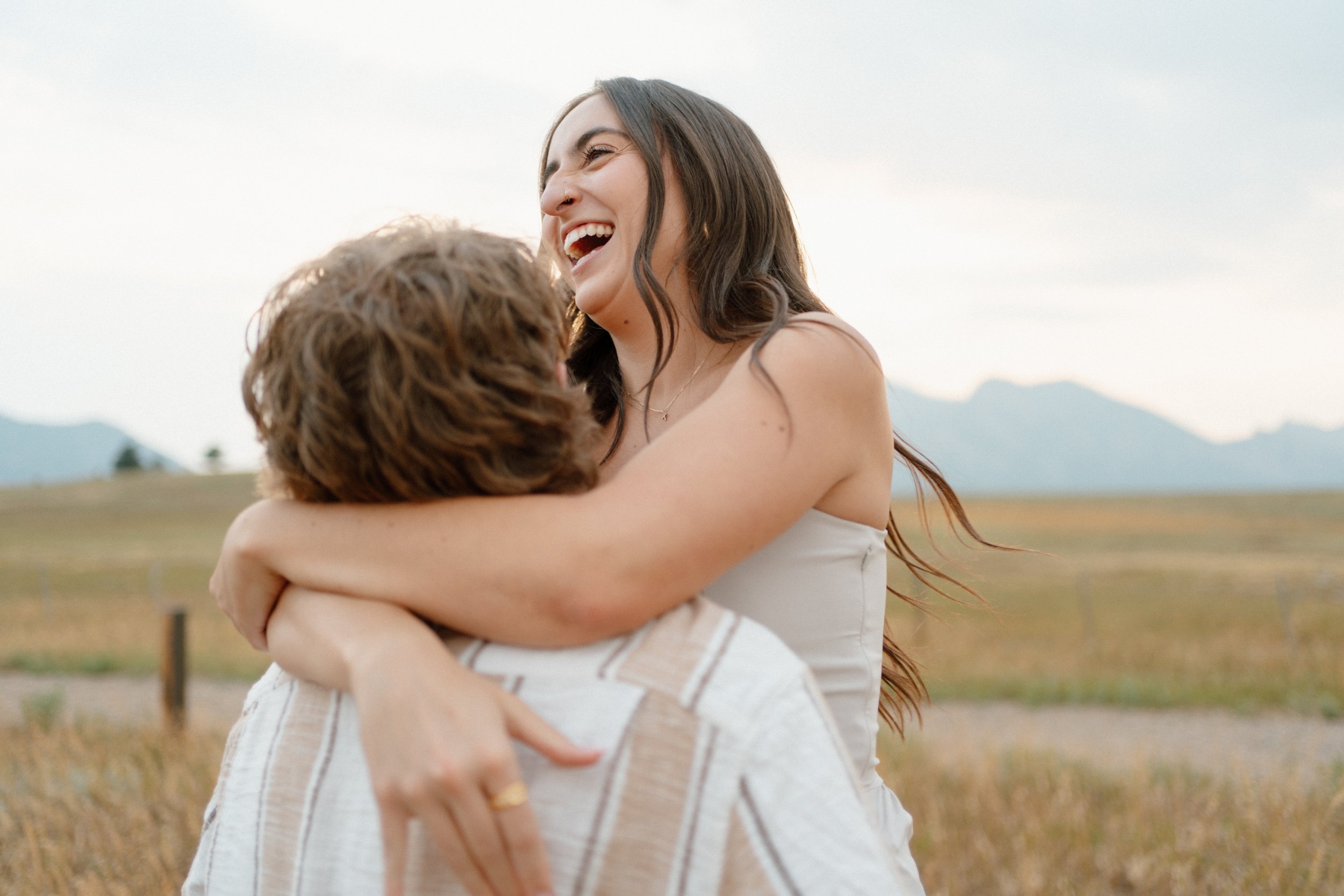 A woman with long dark hair and a white sleeveless dress hugging a man with curly brown hair in an open field with mountains in the background, smiling and laughing.