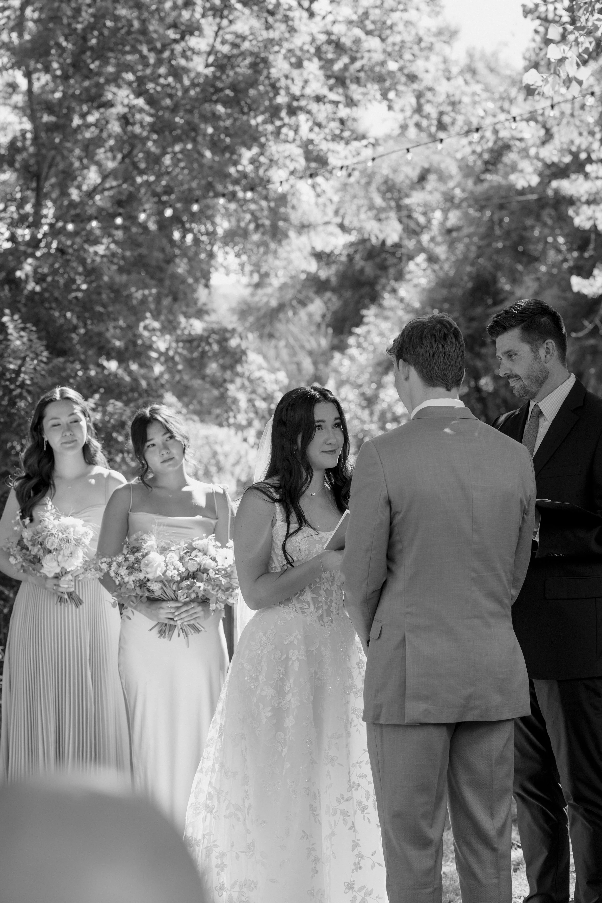 Black-and-white photo of a wedding ceremony outdoors with the bride and groom facing each other, surrounded by bridesmaids and a man officiating.
