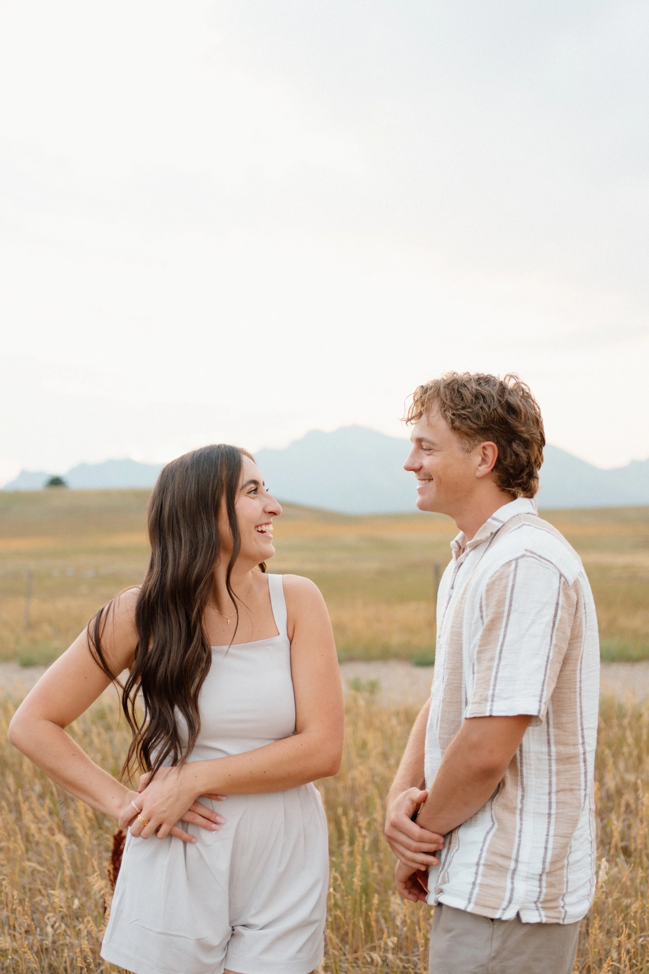 A young woman and man smiling at each other outdoors in a field with mountains in the background.