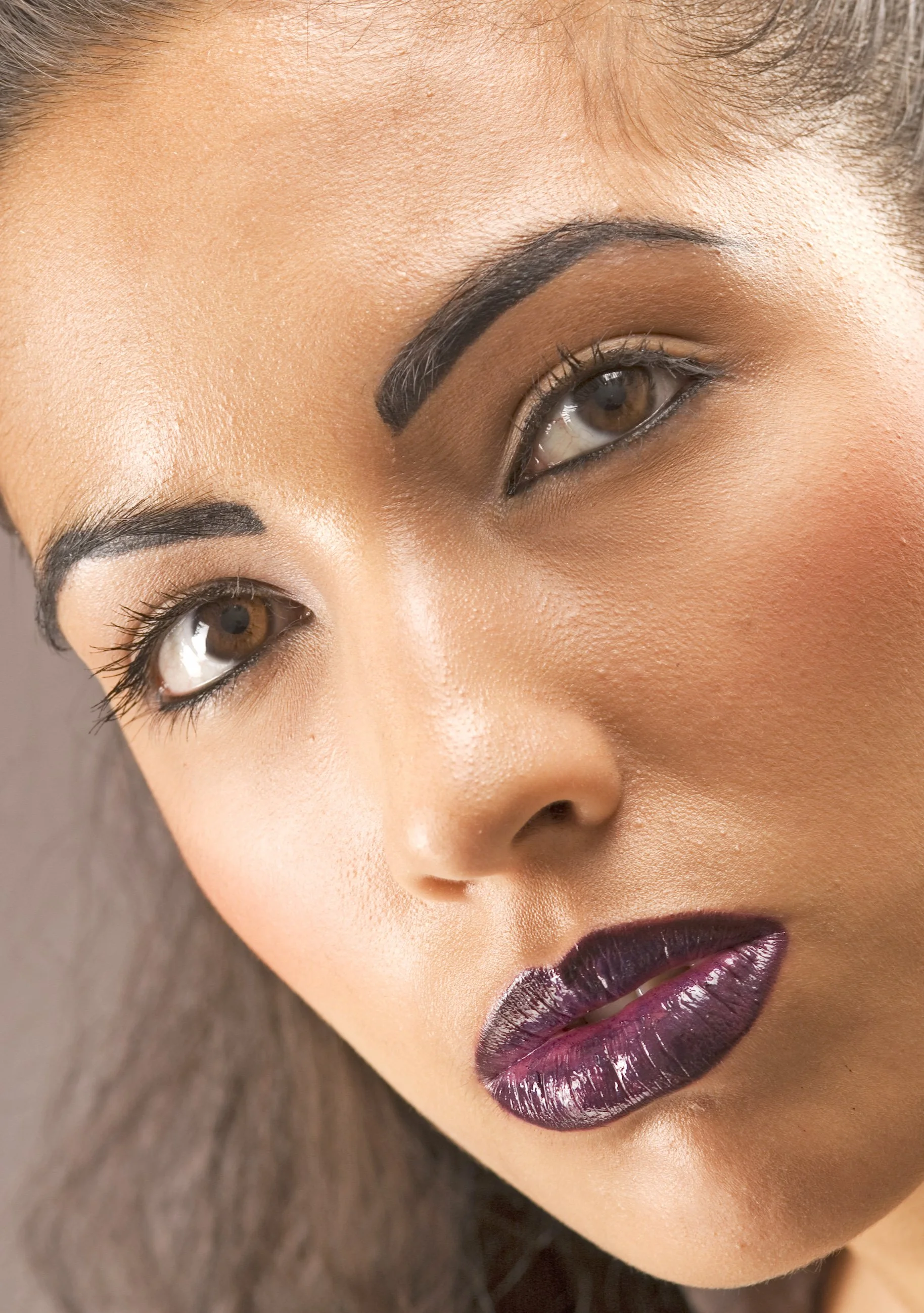 Close-up of a woman's face with makeup, dark eyebrows, brown eyes, and glossy dark purple lipstick.