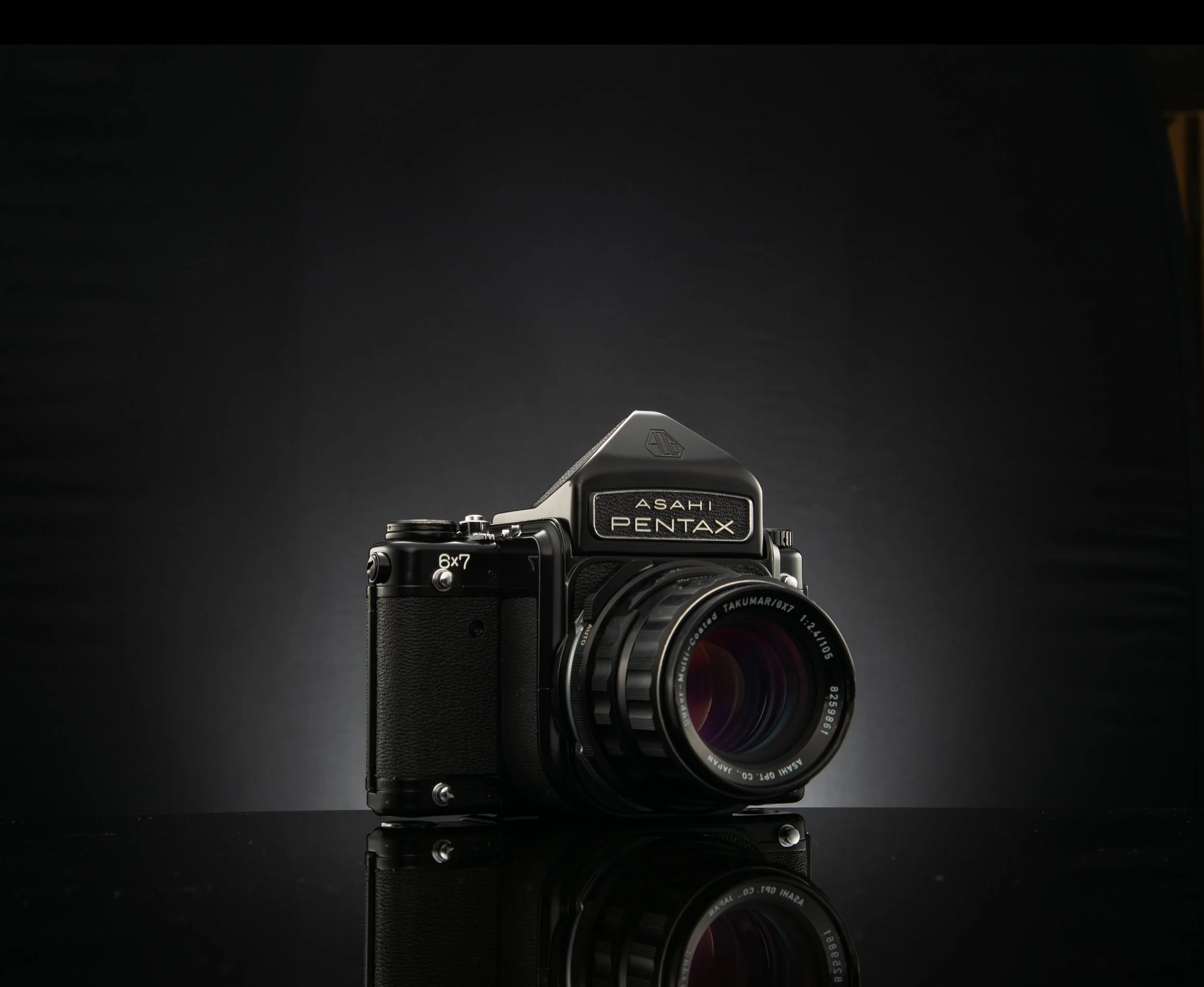 A vintage Asahi Pentax 6x7 camera with a Takumar 105mm lens, placed on a reflective black surface against a dark background.