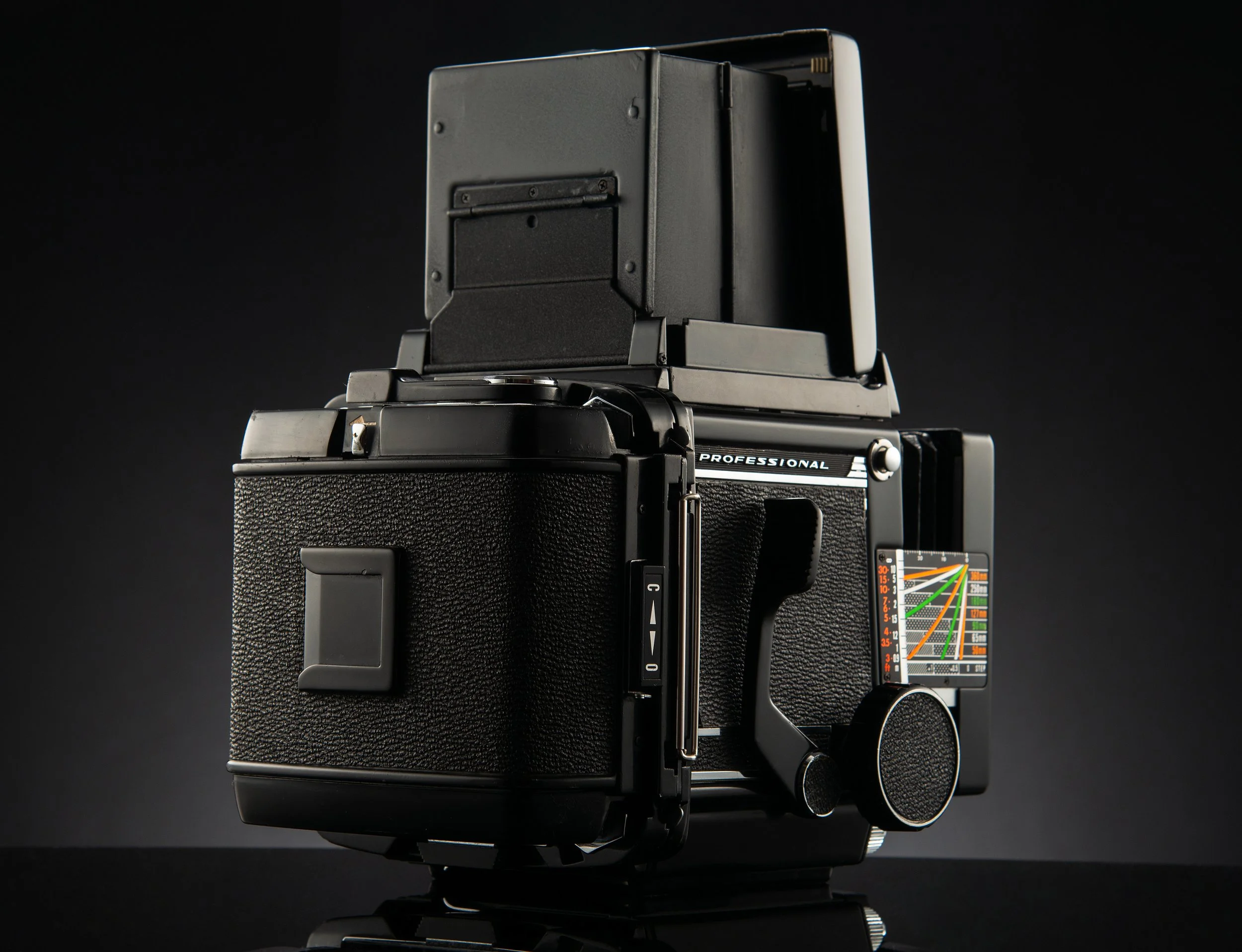 Professional medium format film camera with a viewfinder, textured black body, and various dials and attachments, set against a dark background.