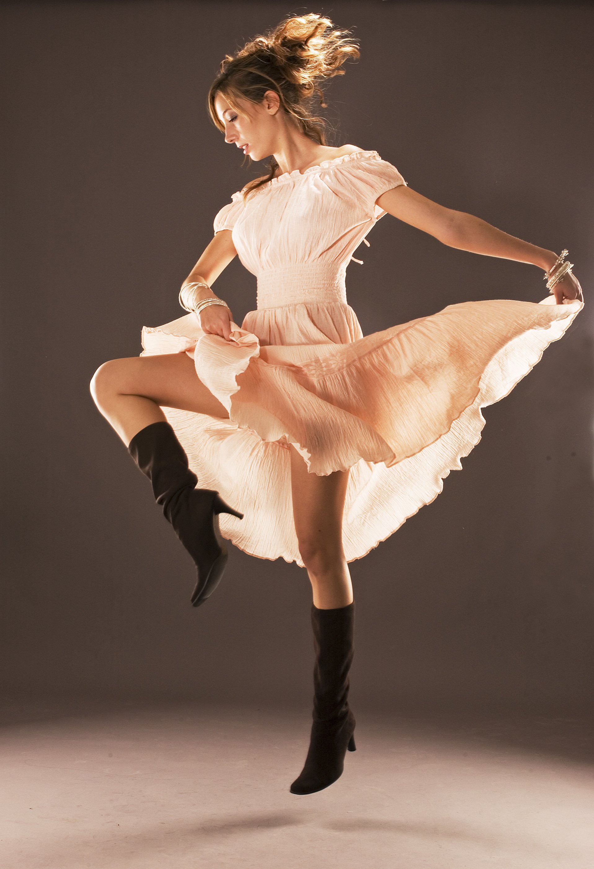 A woman in a peach-colored dress and black knee-high boots jumping in the air against a plain background.