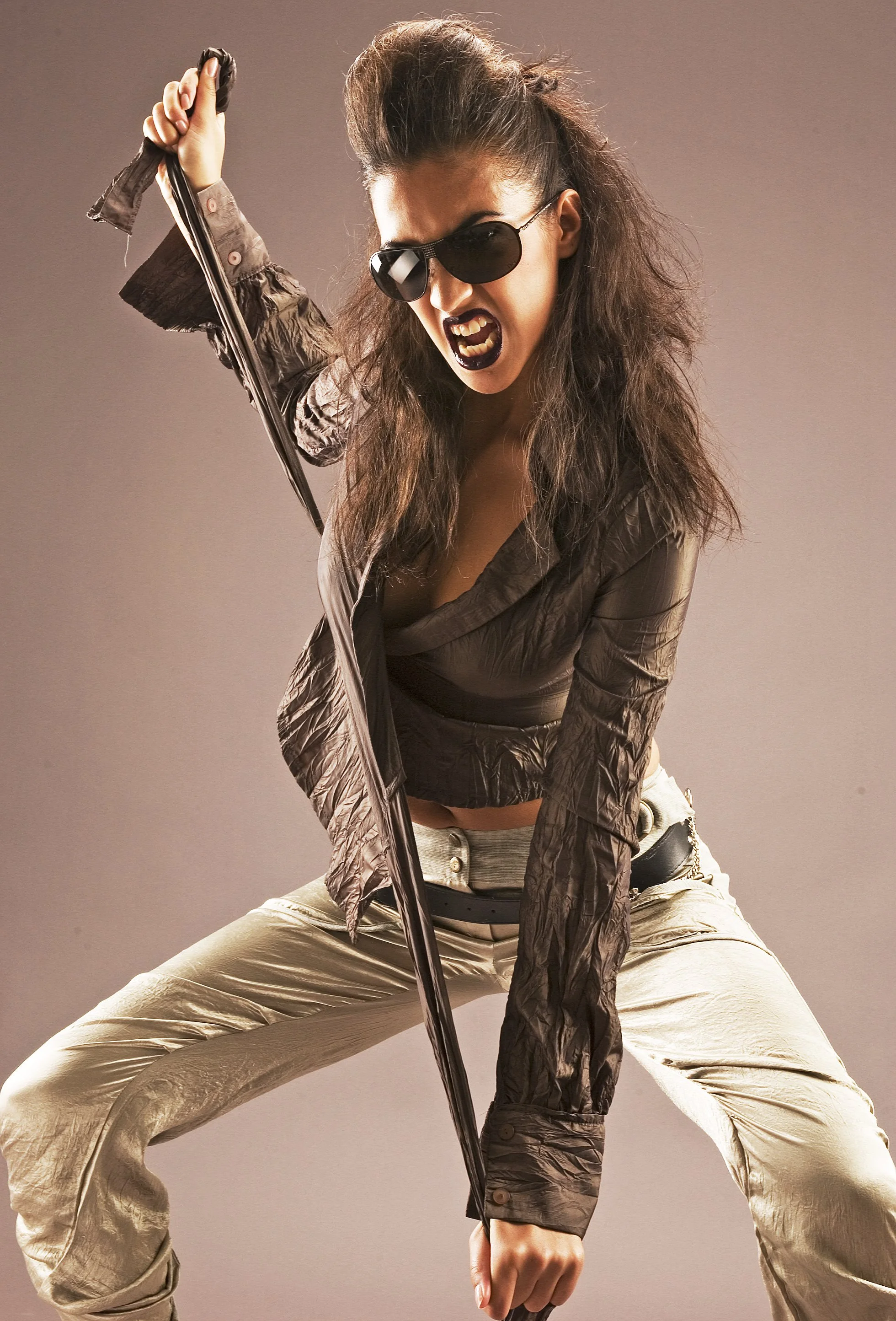 A woman with long, messy hair wearing sunglasses, black lipstick, a dark shiny jacket, and beige pants, is crouching and holding a sword in a fierce pose.
