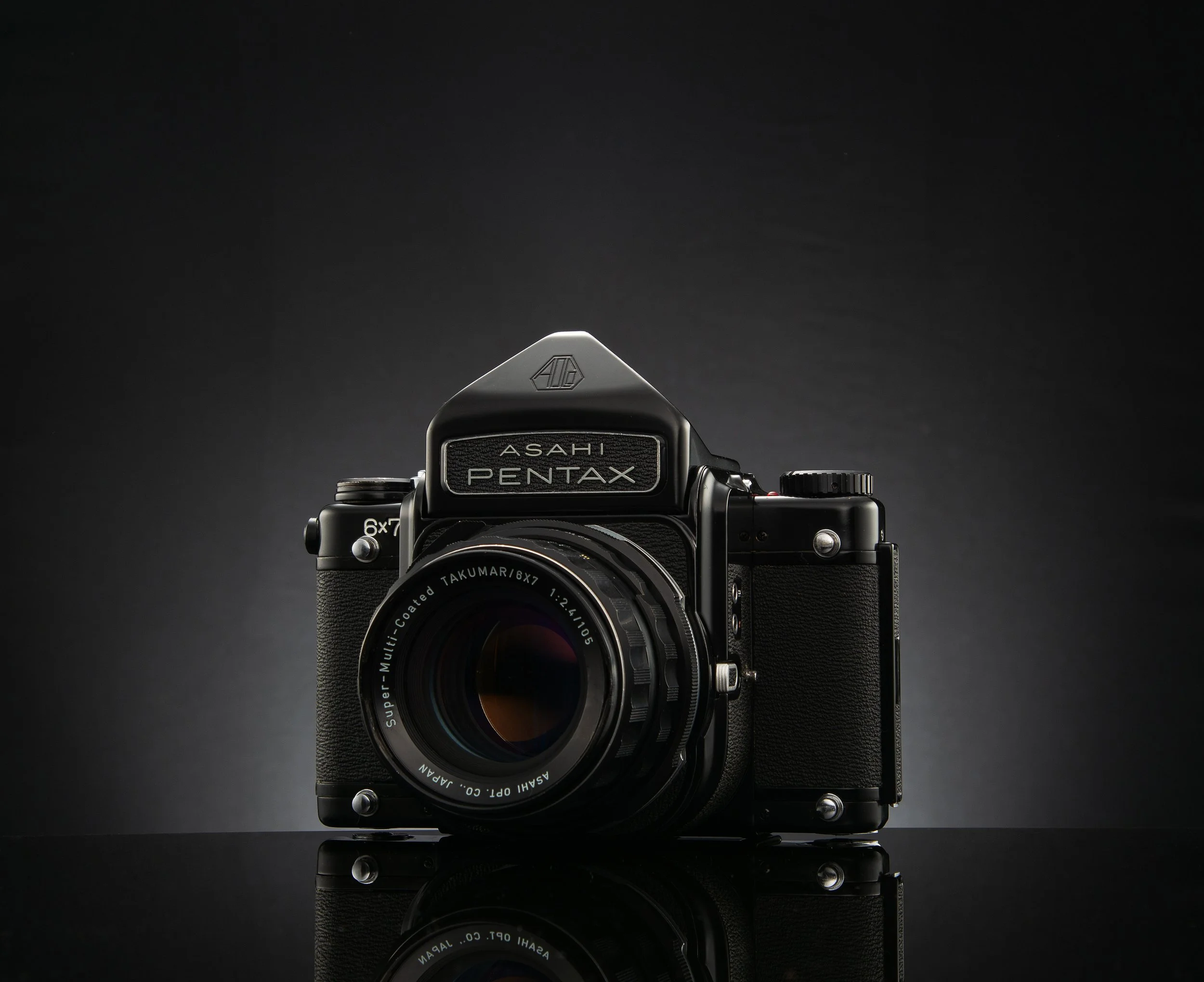 Black vintage Asahi Pentax camera with a large lens on a reflective black surface against a dark background.