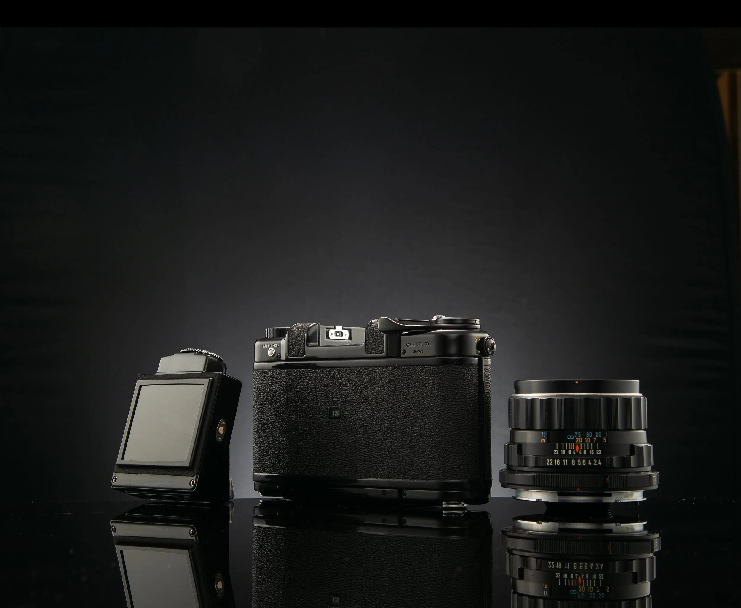 A black vintage camera with multiple lenses, and a small digital camera on a reflective black surface.