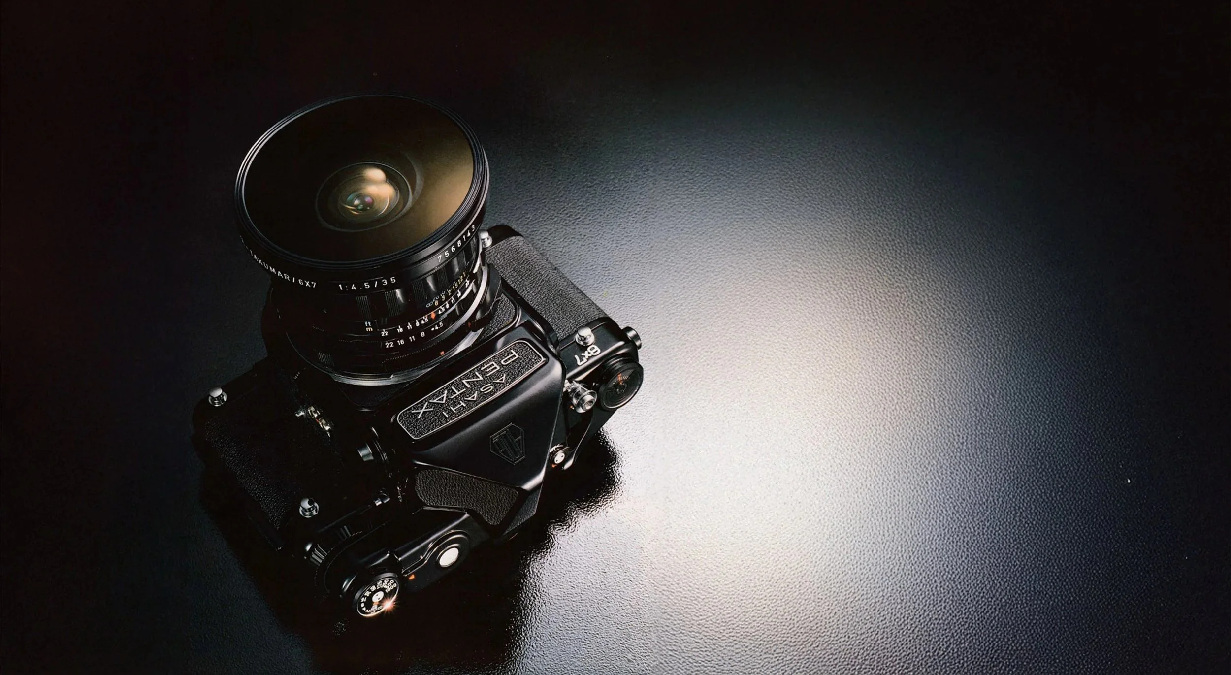 A black camera with a lens attached, placed on a dark textured surface, with a light source illuminating from the right side.