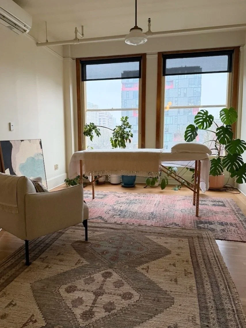 Room with large windows, sofa, plants, and a massage table or bed surrounded by rugs.