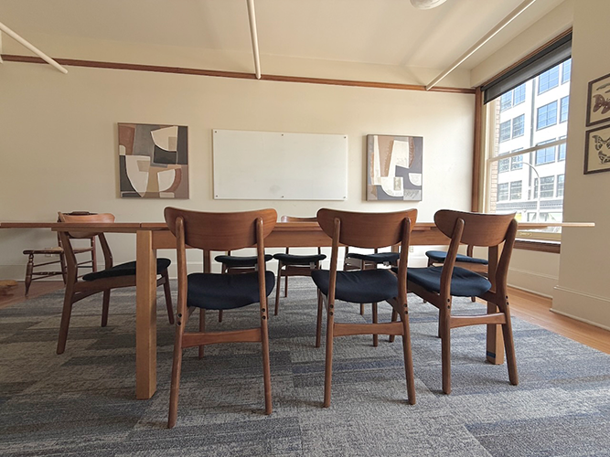 Circle Conference Room has a long table surrounded by mid-century modern chairs. Large windows show downtown Portland, Oregon. The wall behind the table has three abstract art pieces and a whiteboard. There is a flat-screen TV on the other wall.