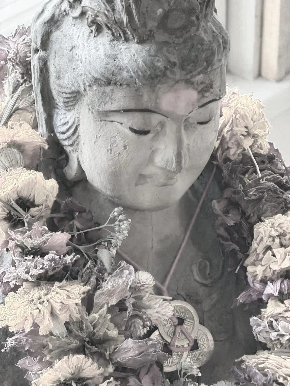 A close-up of a Buddha statue's face surrounded by dried flowers and traditional Chinese coins on a string.