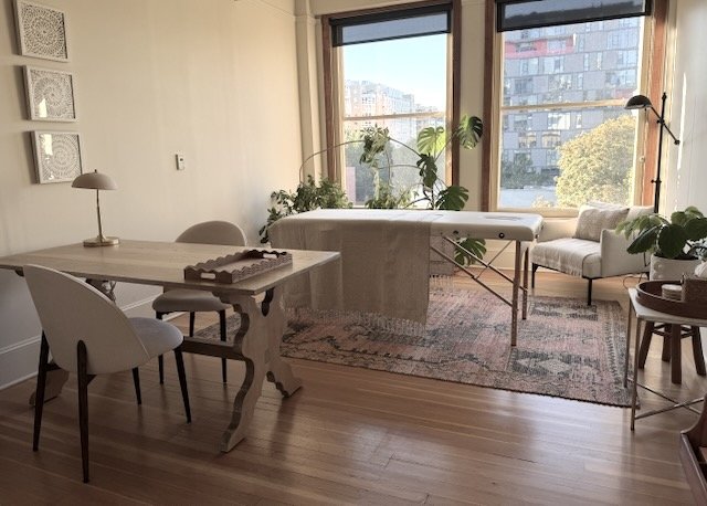 Prism Studio is a bright treatment room and private office with large windows, plants, a massage table, a desk with chairs, and a cozy armchair, decorated in neutral earthy tones.